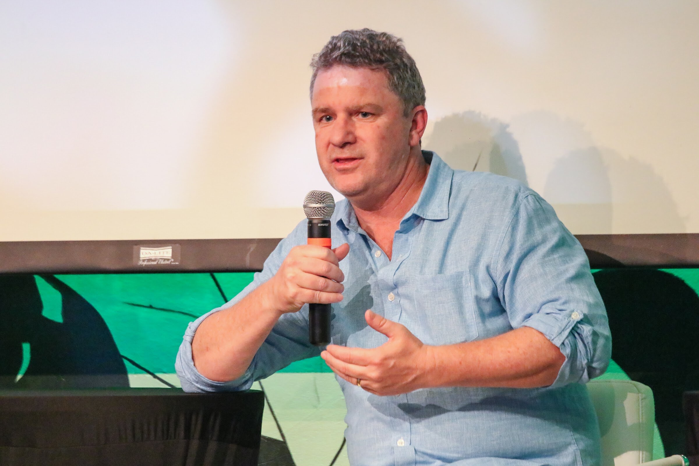 A man in a rolled-sleeve light blue shirt holds a microphone and gestures with one hand against a white screen and green wall art.