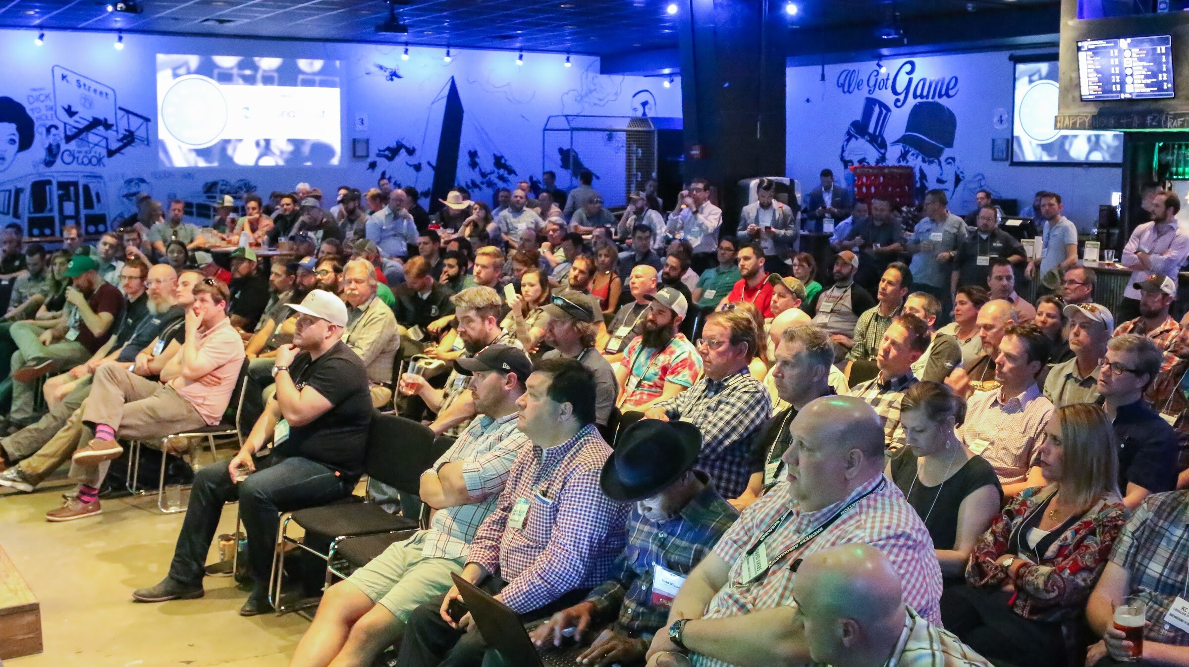 Rows of seated attendees with lanyards watch the stage, with blue-lit murals and TV screens on the wall behind them.