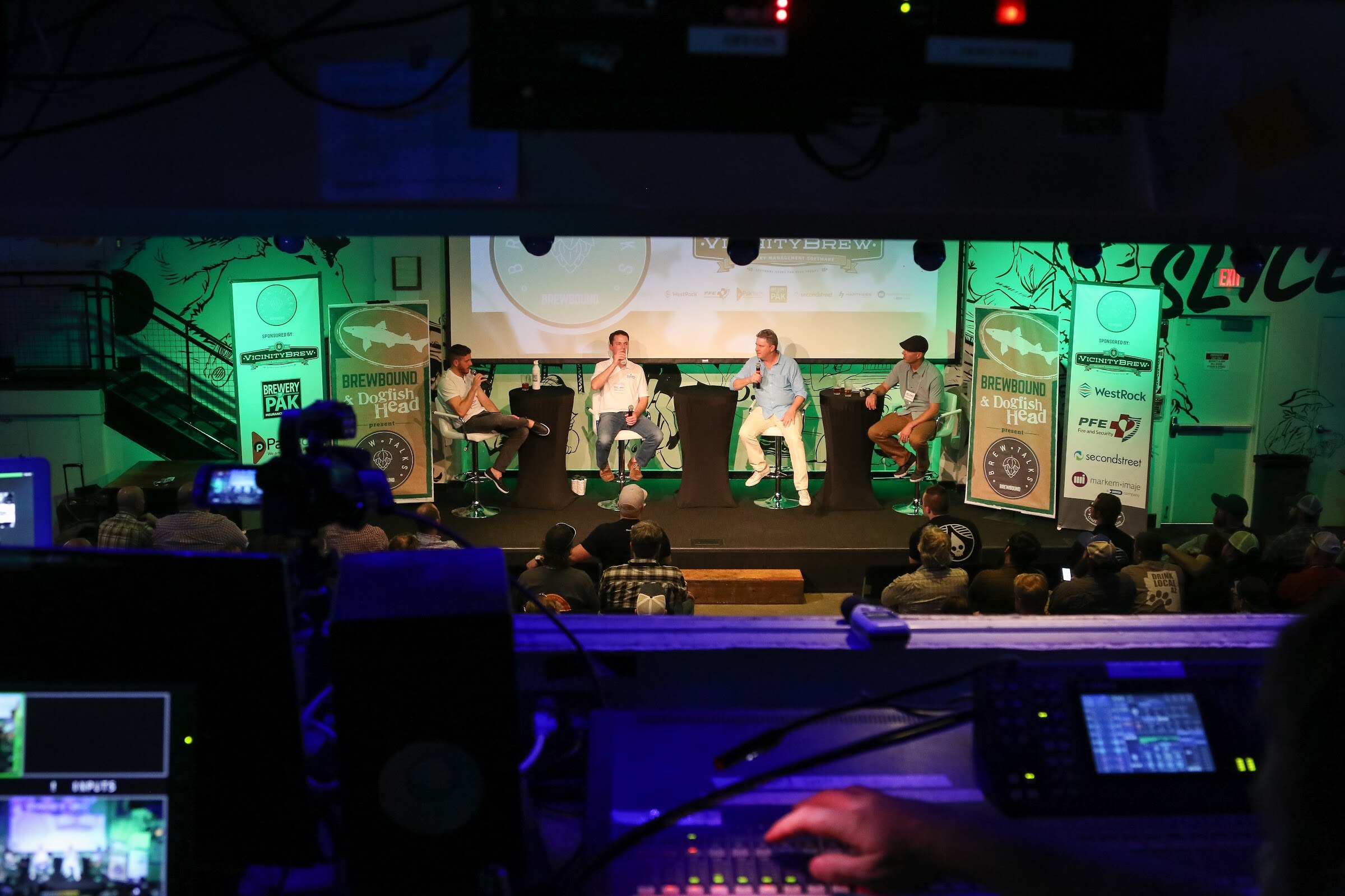 A dim view over audio equipment and a hand on a mixing console toward a lit stage with four seated panelists and sponsor banners.