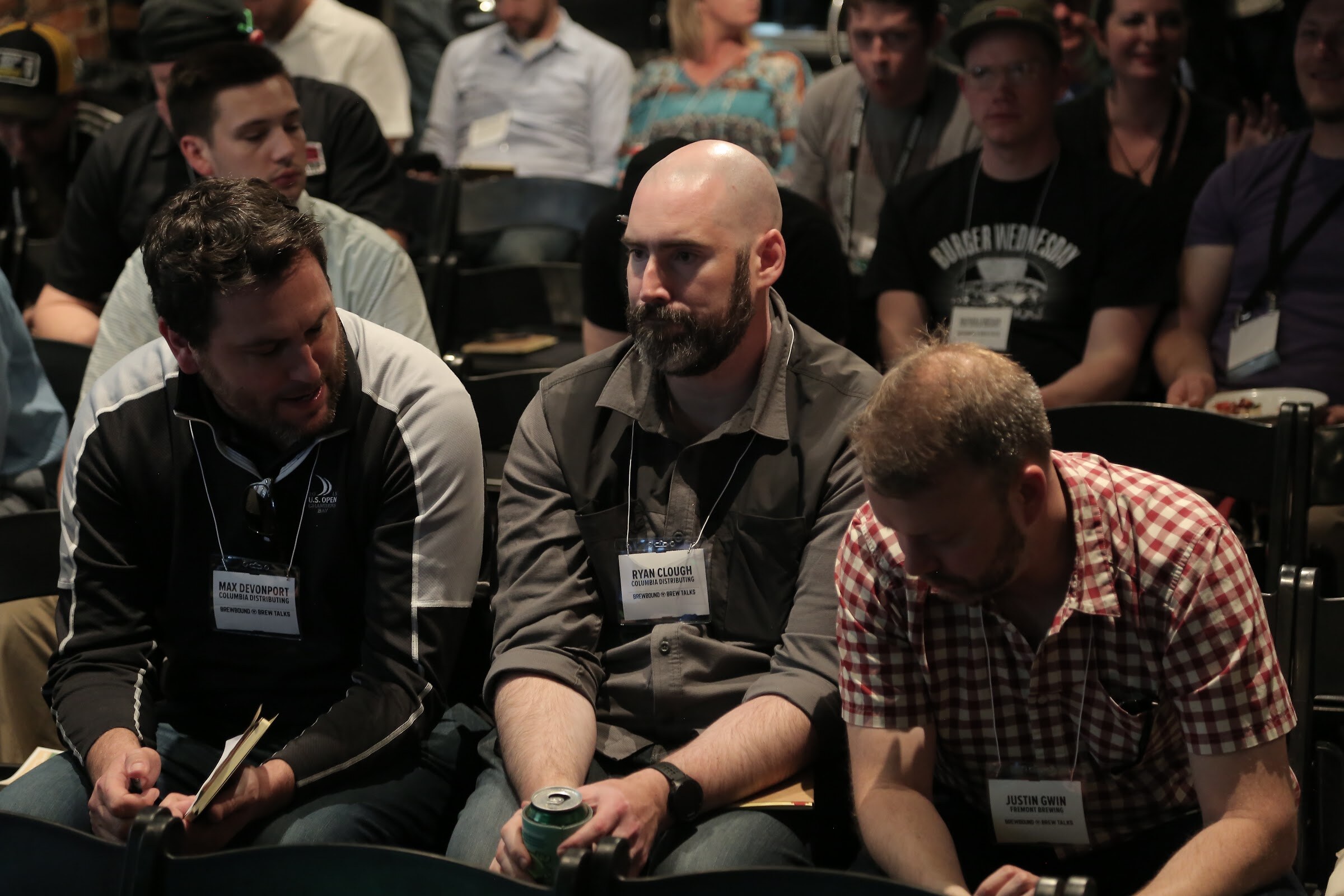 Three badge-wearing men sit close together in folding chairs, one holding a can, with more attendees in rows behind them.