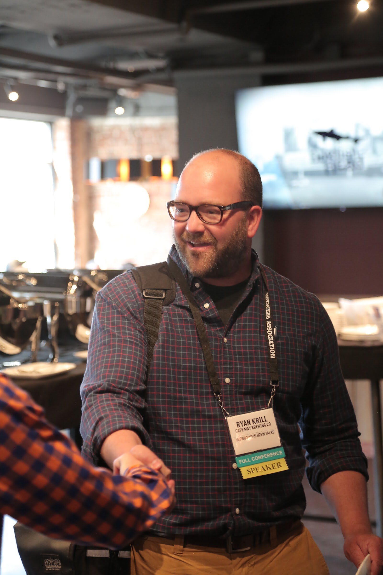 A bald, bearded man in glasses and a plaid shirt with a lanyard shakes hands with someone at the edge of the frame.