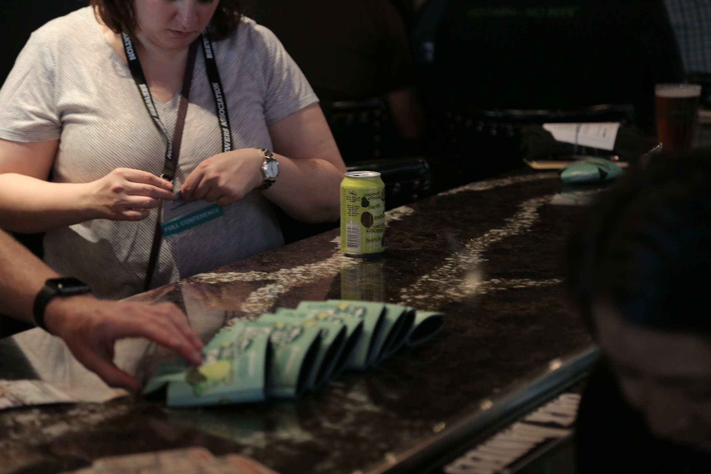 A woman looks down at her lanyard badge beside a green beer can and a row of mint-green koozies on a dark granite counter.