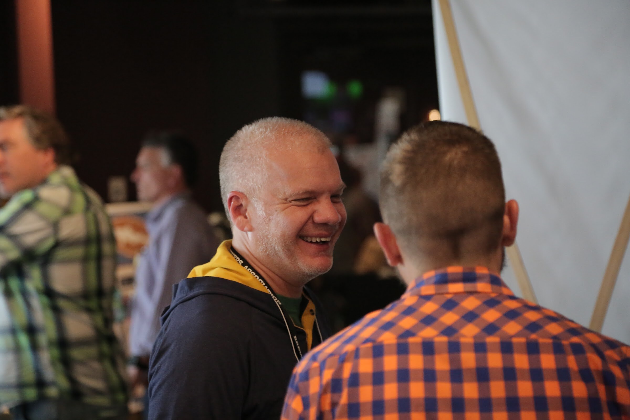 A man in a navy and yellow hoodie laughs while talking with a man in an orange check shirt seen from behind.
