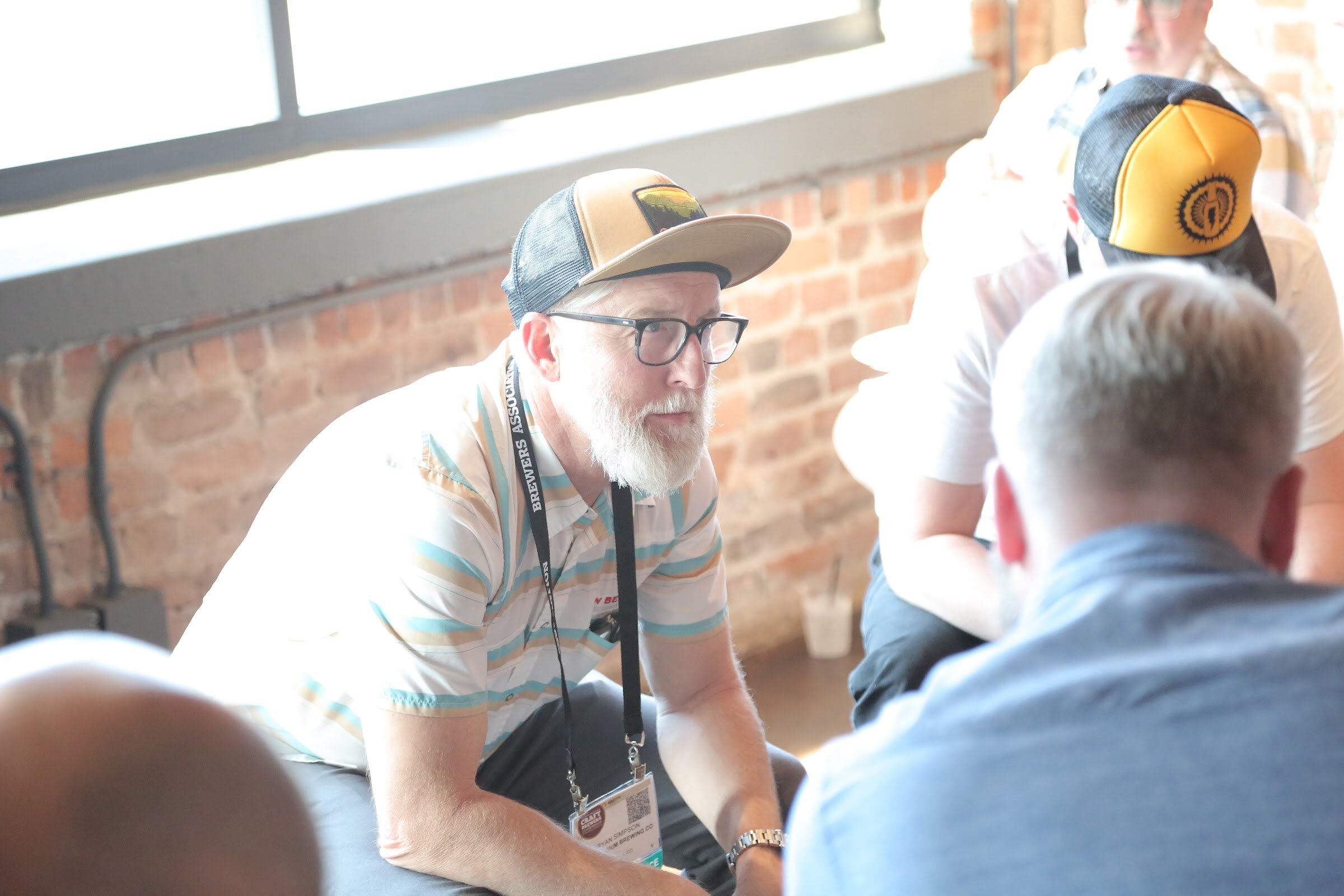 A white-bearded man in a striped polo, glasses and a snapback cap sits forward in front of a brick wall, listening to two other people.