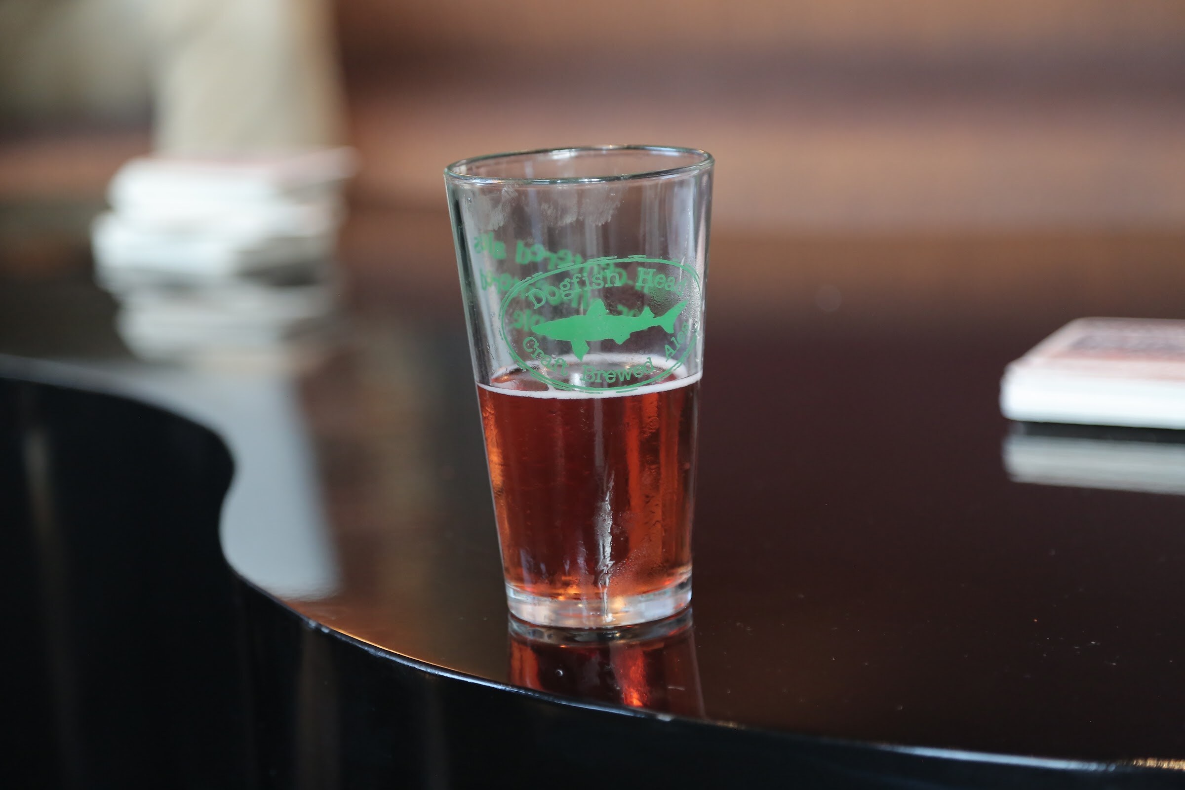 A pint glass with a green Dogfish Head logo half full of amber beer on a glossy black surface, with coasters blurred behind.