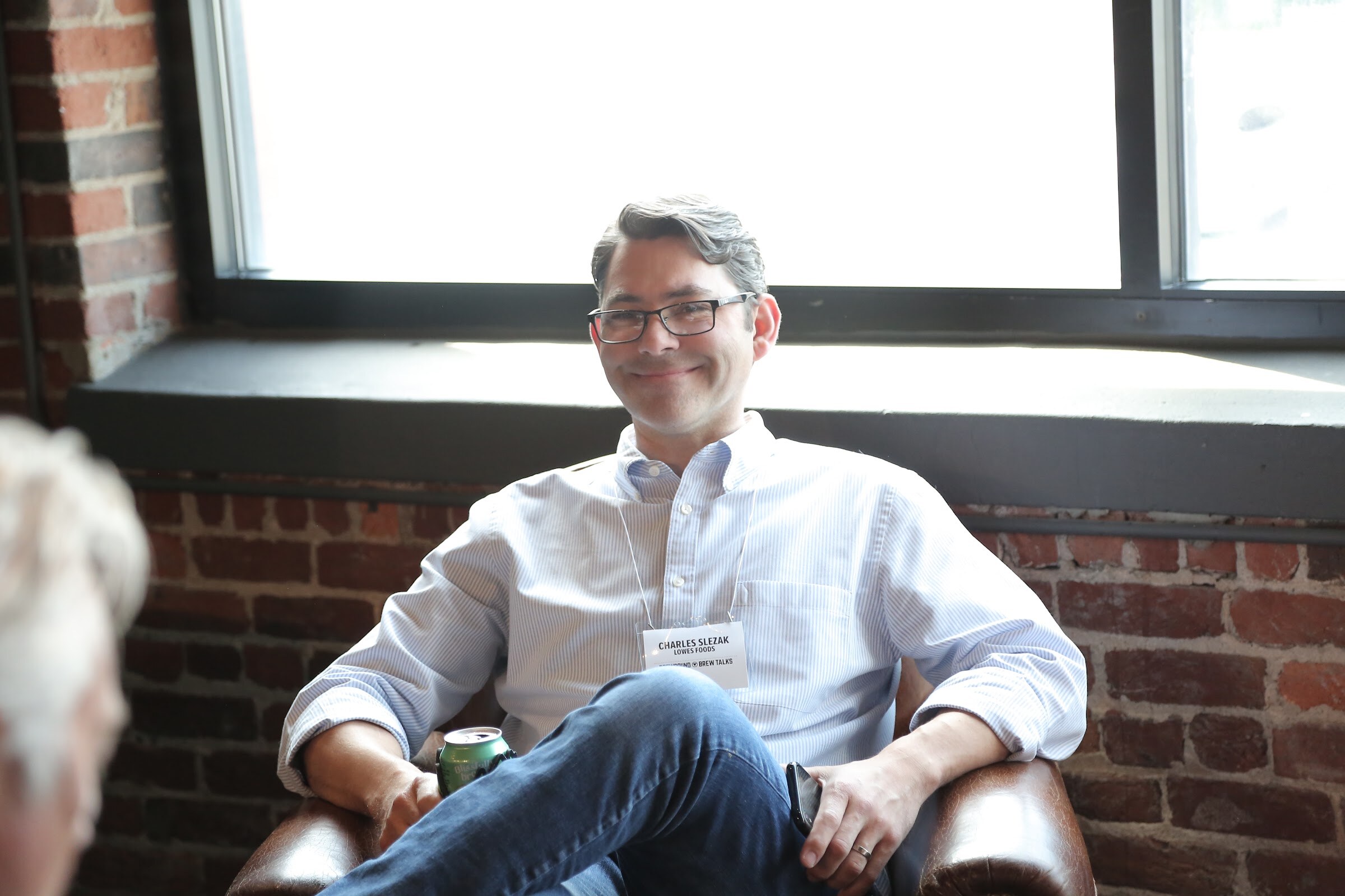 A man in glasses and a light button-down shirt sits back in a brown leather chair in front of a brick wall and a large window.