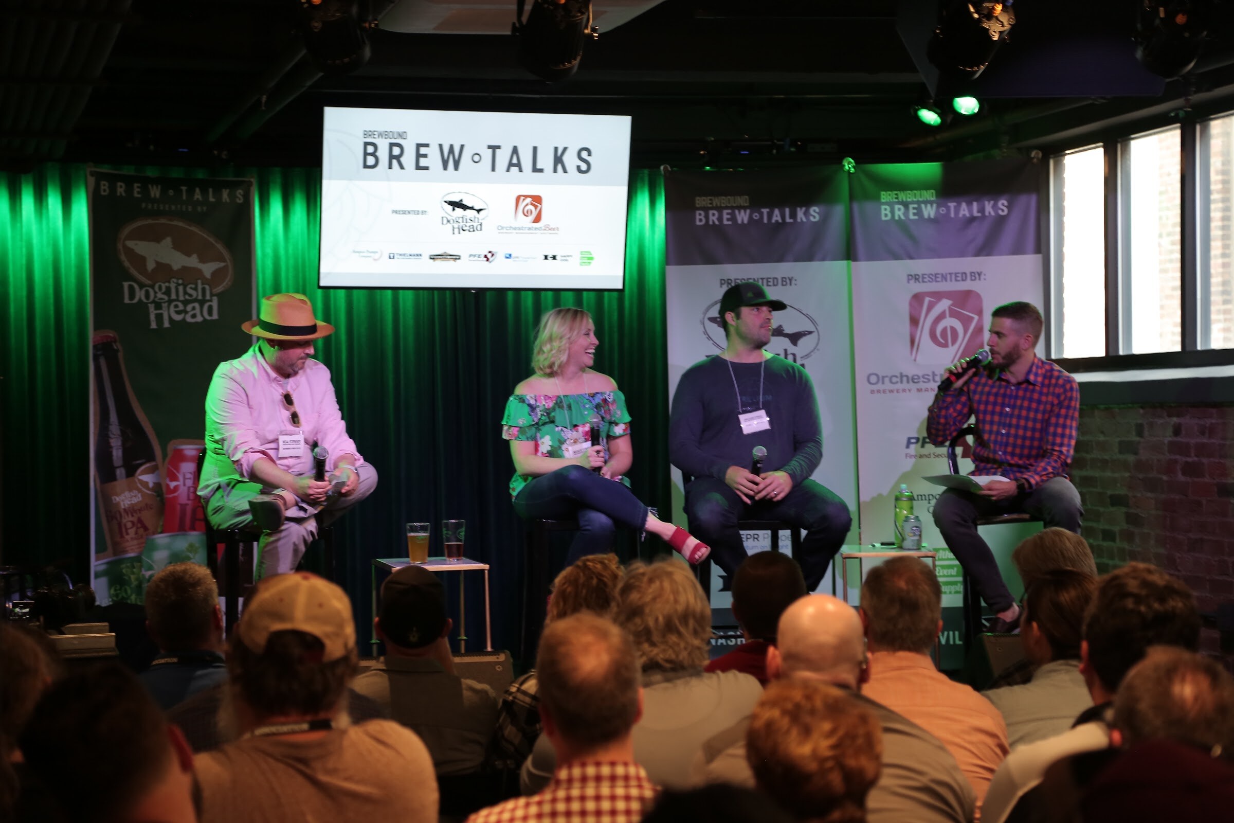 Three panelists and a moderator on stools in front of green curtains, a Brew Talks screen and banners for Dogfish Head and Orchestrated Beer, with the audience in the foreground.
