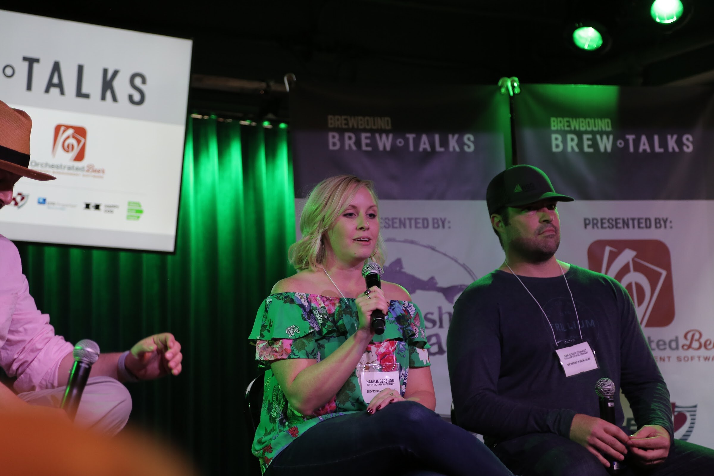 A woman in a green floral top holding a microphone and a man in a black cap sit in front of Brewbound Brew Talks banners with an Orchestrated Beer sponsor logo.