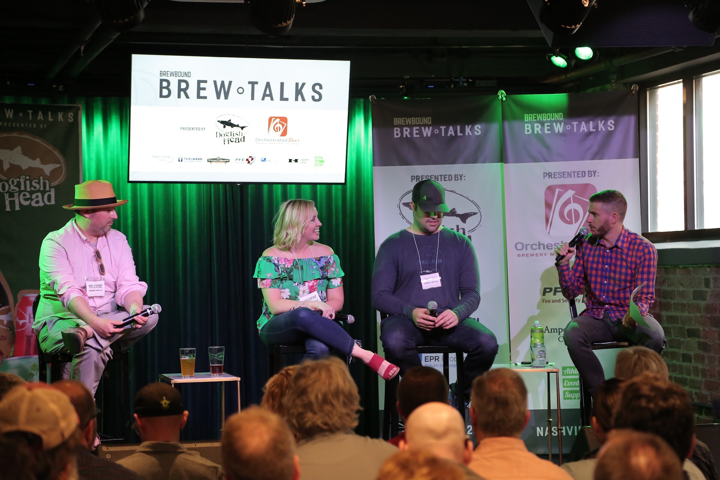 A moderator in a plaid shirt with a microphone and three seated panelists in front of the Brew Talks screen, with the backs of audience heads in the foreground.