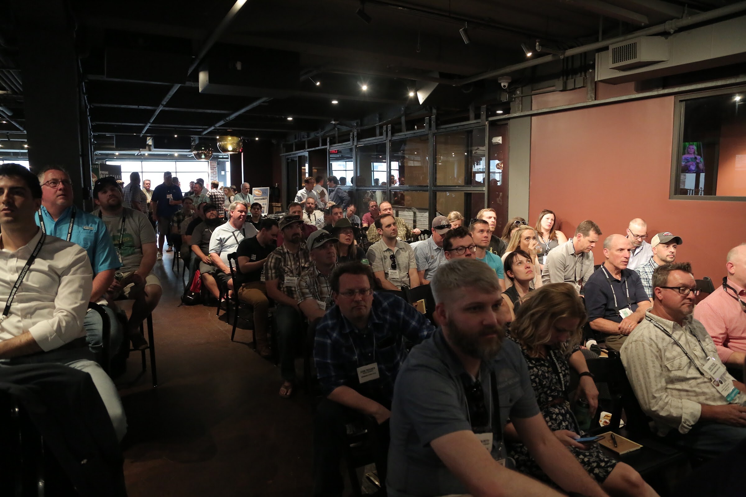 Rows of seated attendees with badges fill the room, with more people standing at the back and a window wall behind them.