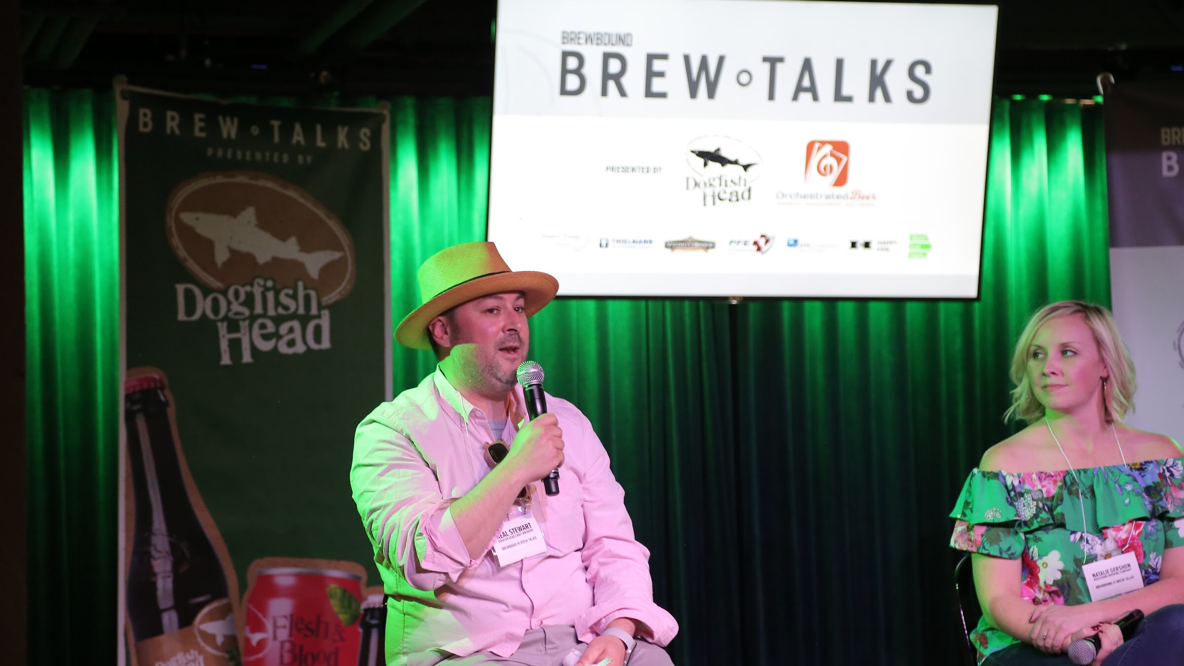 A man in a straw hat and light pink shirt holds a microphone in front of green curtains, a Brew Talks screen and a Dogfish Head banner with a Flesh & Blood can.