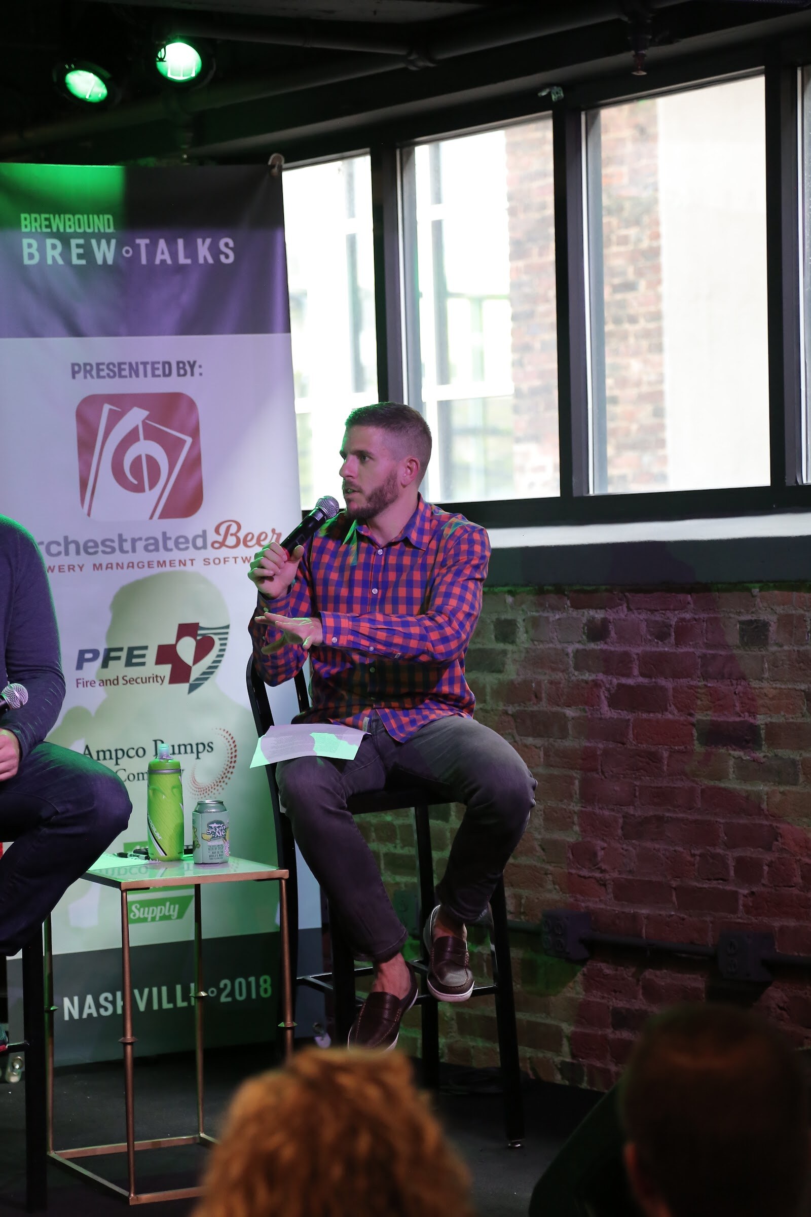 A bearded man in a plaid shirt speaks into a microphone on a tall stool beside a banner reading Presented By Orchestrated Beer, with NASHVILLE 2018 along the bottom.