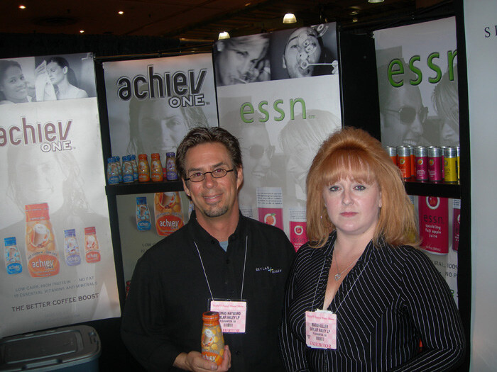 Thomas Hayward, left, holds an achiev ONE bottle beside Maggi Keller of Skylar Haley LP, in front of achiev ONE and essn banners and a shelf of essn cans.