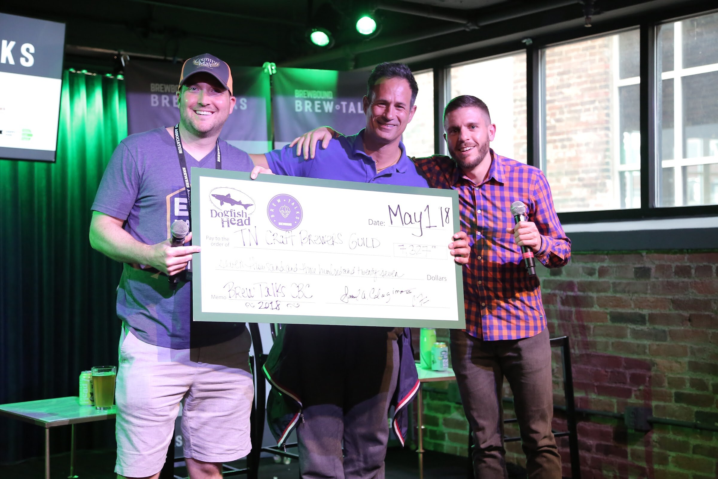 Three smiling men with microphones stand on stage holding a large cardboard check with Dogfish Head and Brew Talks logos dated May 1, 2018.