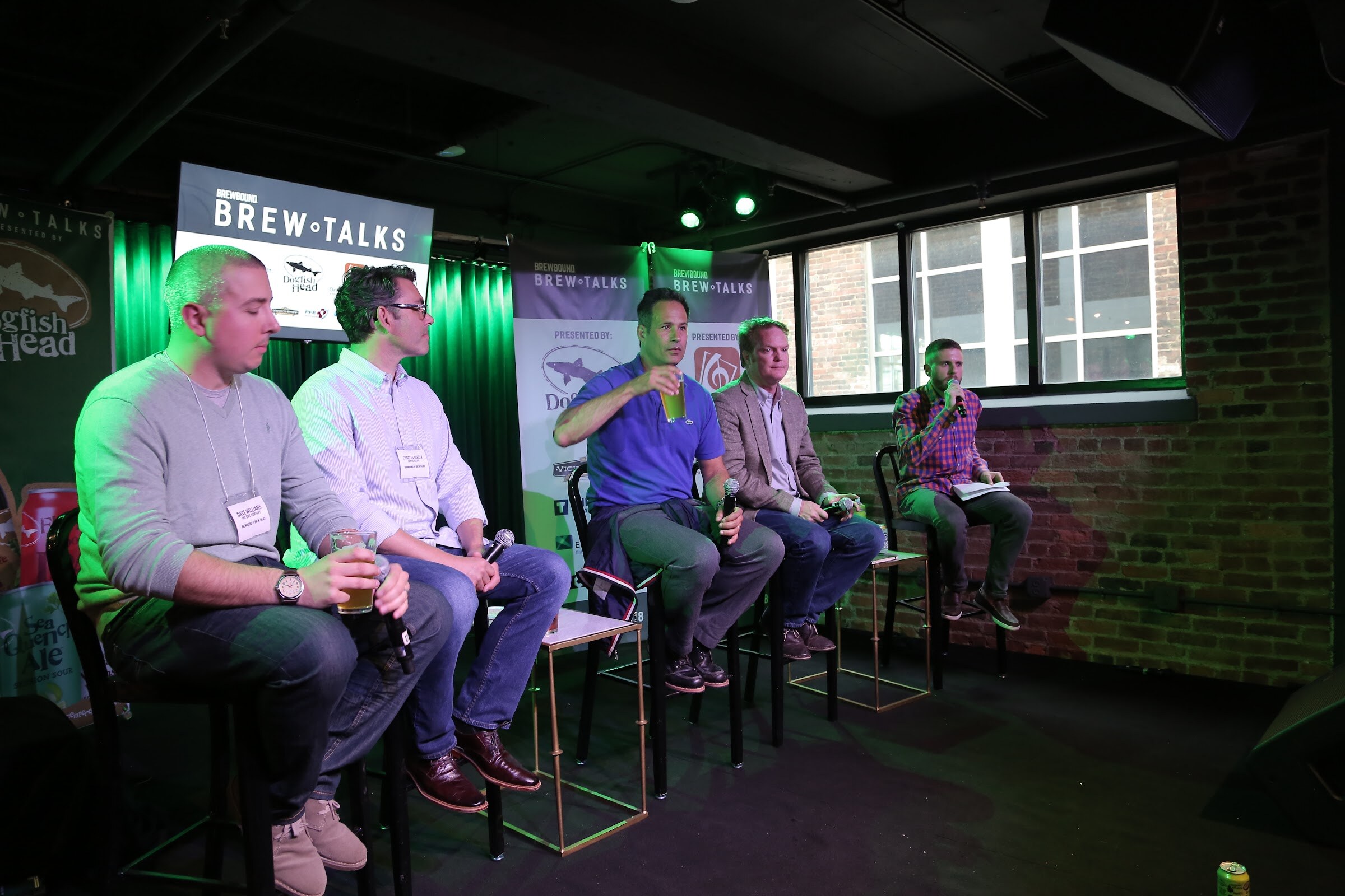 Four men holding drinks and microphones on bar stools face a moderator in a plaid shirt, with a Brew Talks screen and banners behind them.
