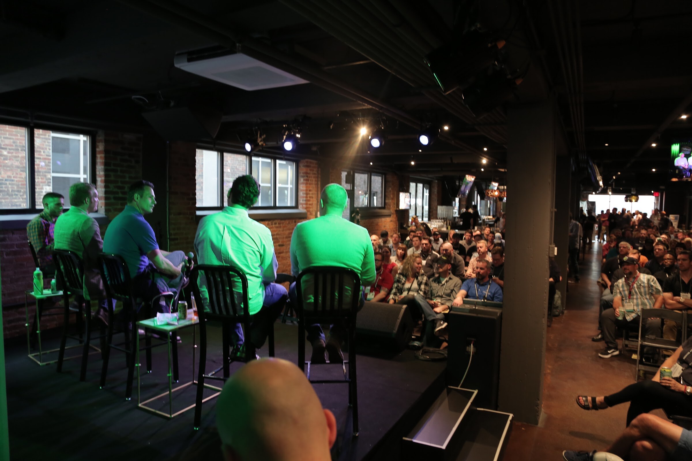 Five people seated on stools on a stage in a brick-walled bar, with a crowd of attendees seated and standing in the rows to the right.