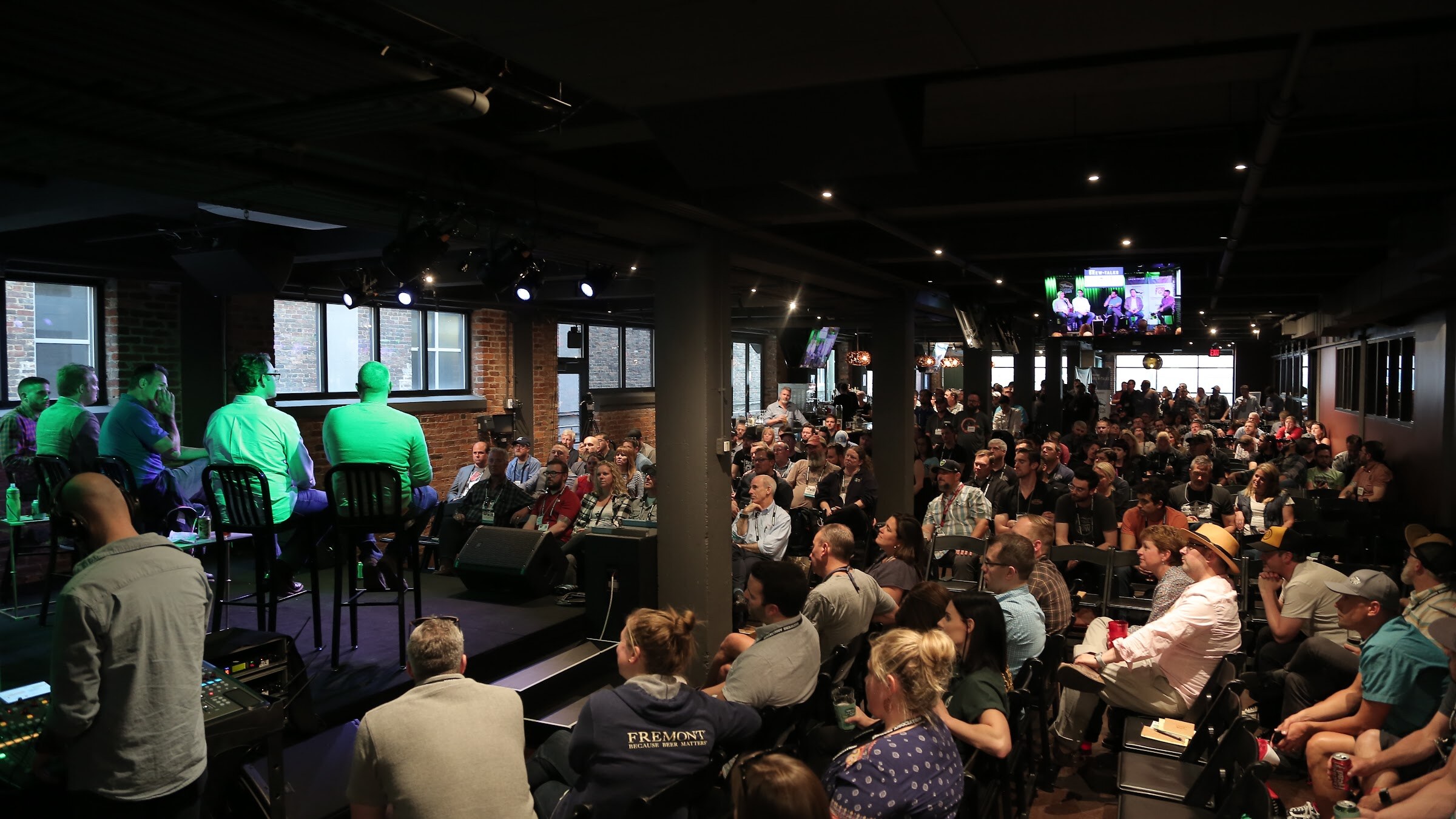 A wide view of a dark brick room filled with attendees, with the panelists on stools at left and a screen showing the stage at the back.
