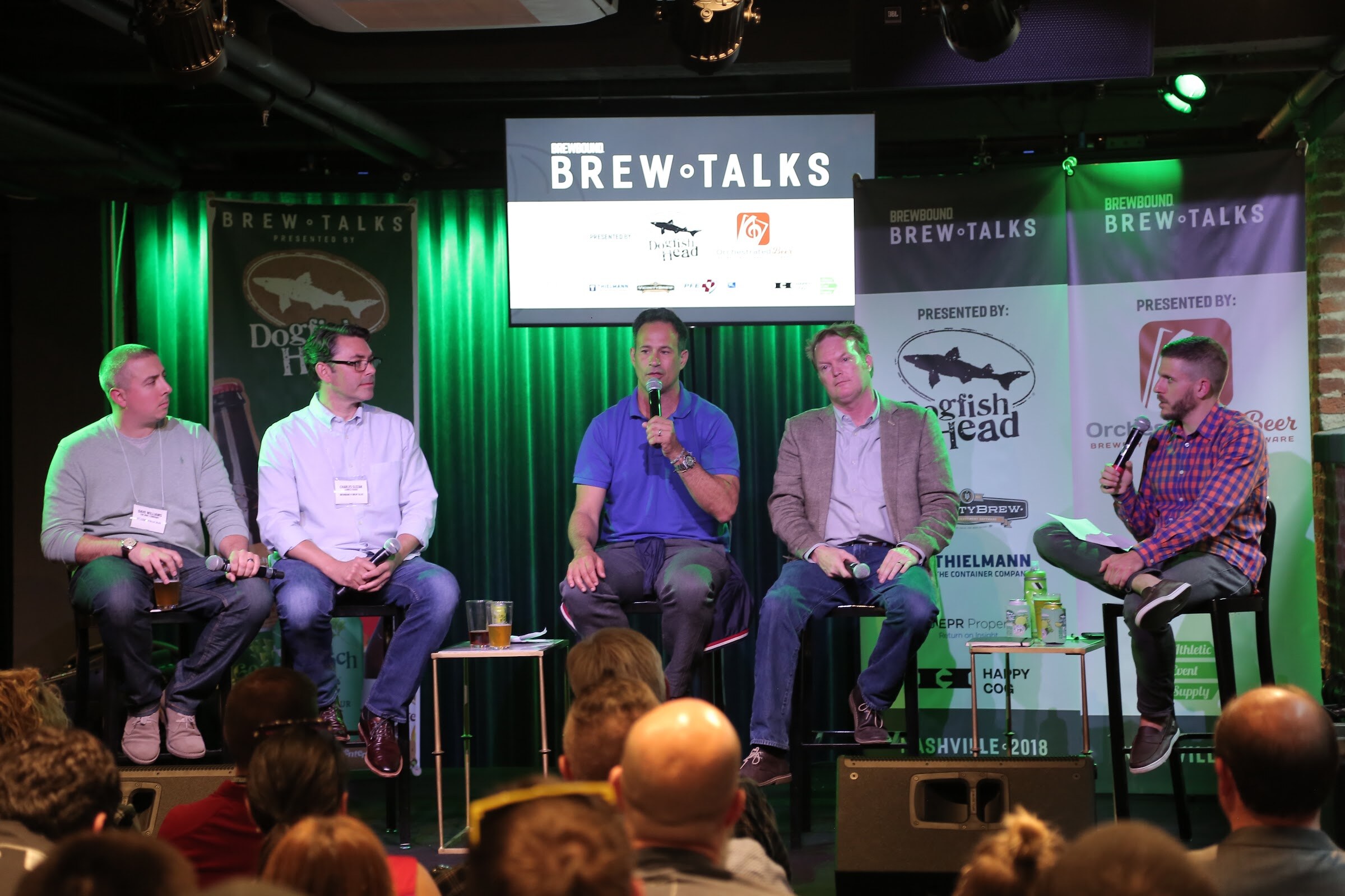 Four men with microphones and drinks on stools, and a moderator in plaid at right, in front of Dogfish Head, Orchestrated Beer, Thielmann and Happy Cog banners.