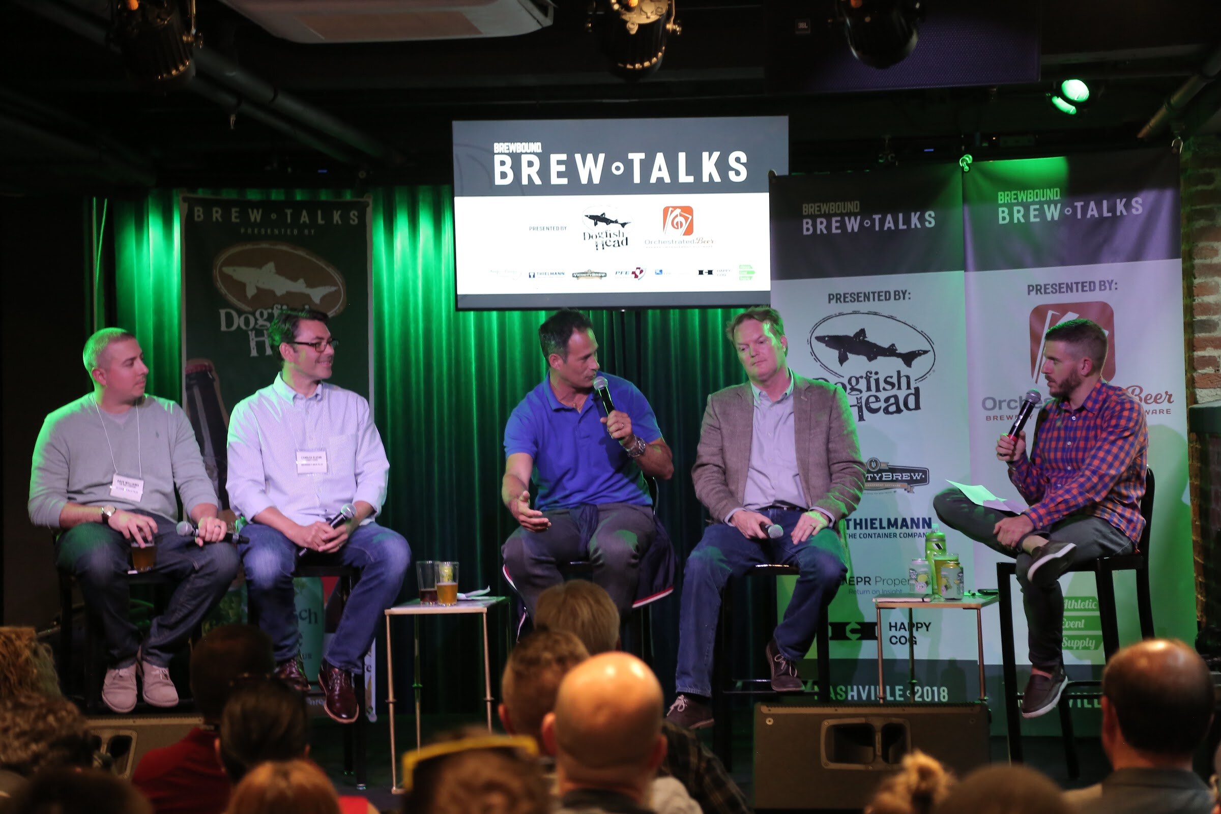 A man in a blue polo speaking into a microphone between four other people on stools in front of green curtains and the Brew Talks screen.