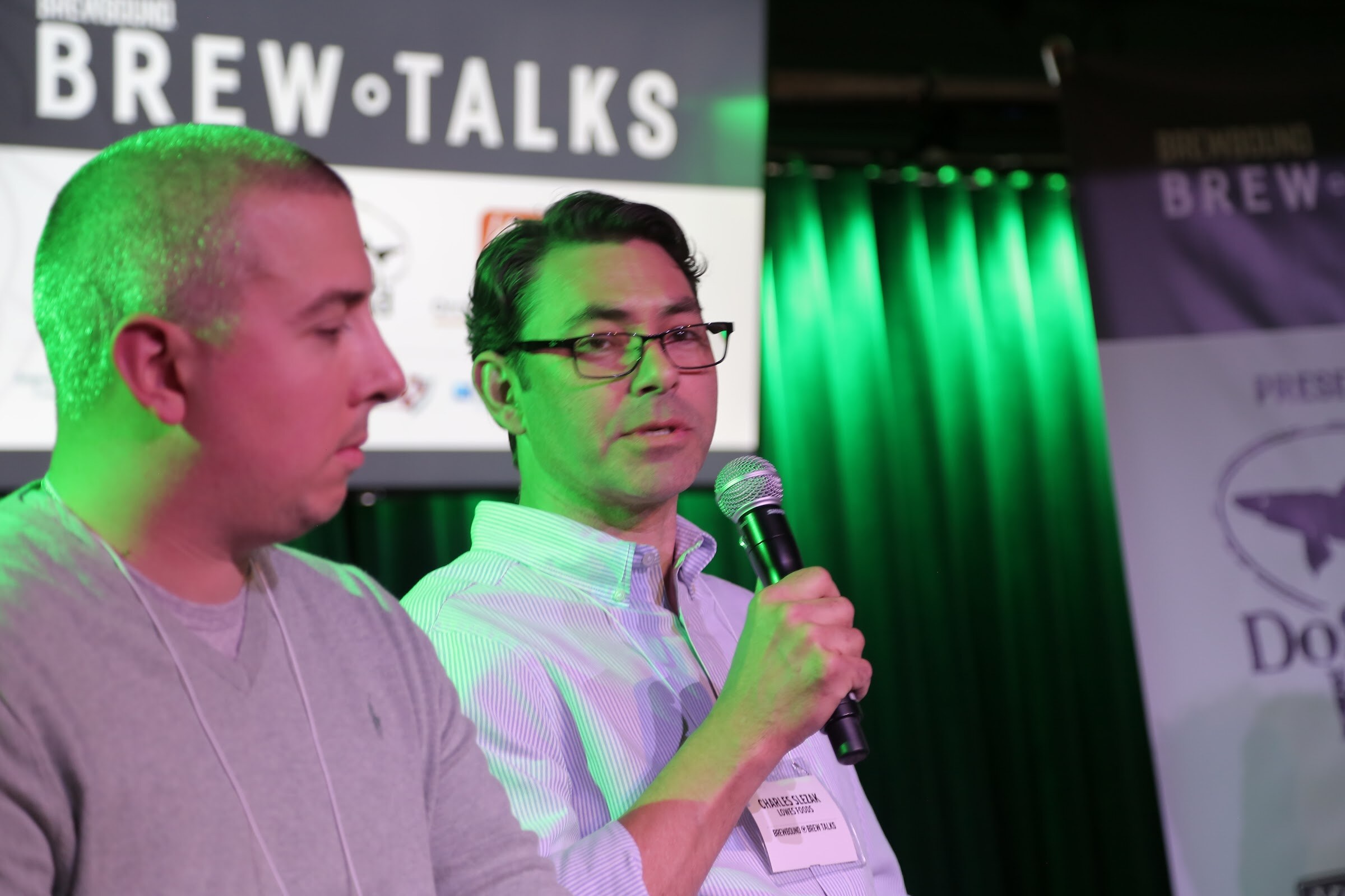 Close view of a man in glasses holding a microphone in front of the Brew Talks screen and green curtains, next to a second man in a gray sweater.