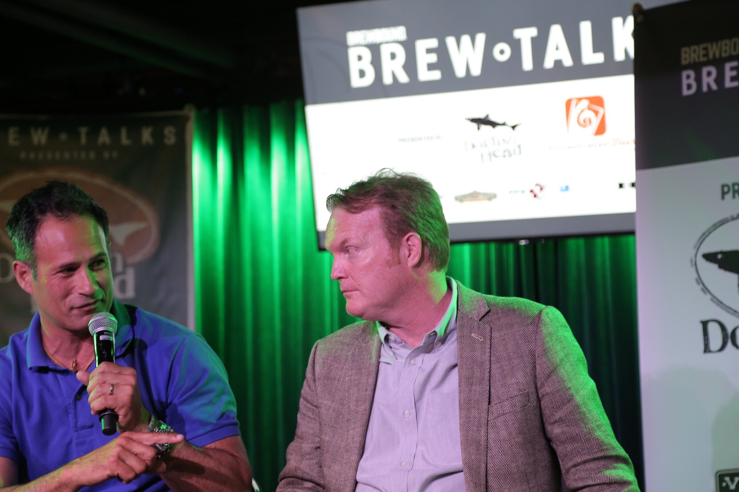 A man in a blue polo with a microphone and a man in a tweed blazer look at one another in front of the Brew Talks screen and green curtains.
