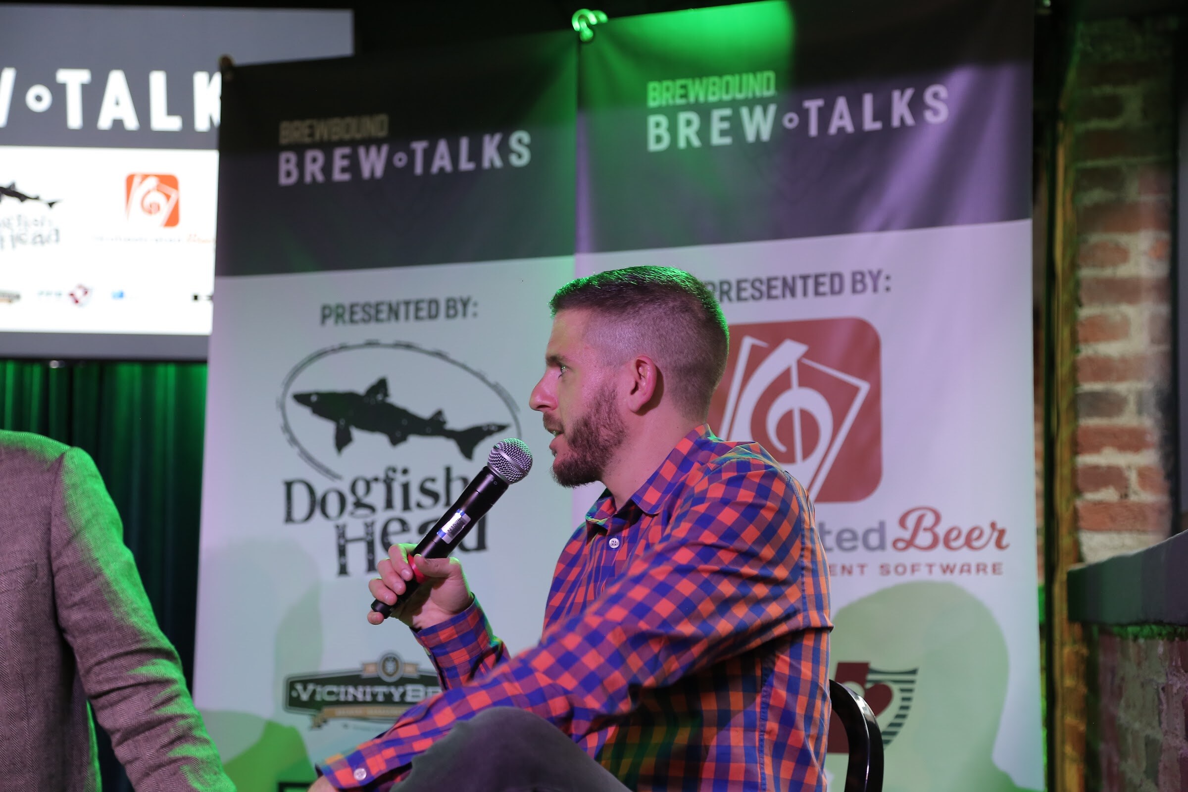 A bearded man in an orange and blue plaid shirt holds a microphone in front of a Brewbound Brew Talks banner showing Dogfish Head, Orchestrated Beer and Vicinity Brewing logos.