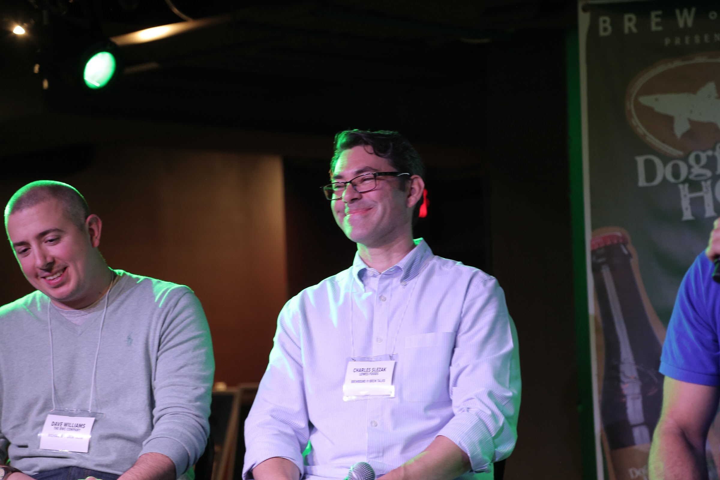 Two men seated side by side on stage with name badges, one looking down and smiling and one smiling broadly, in front of a Dogfish Head banner.