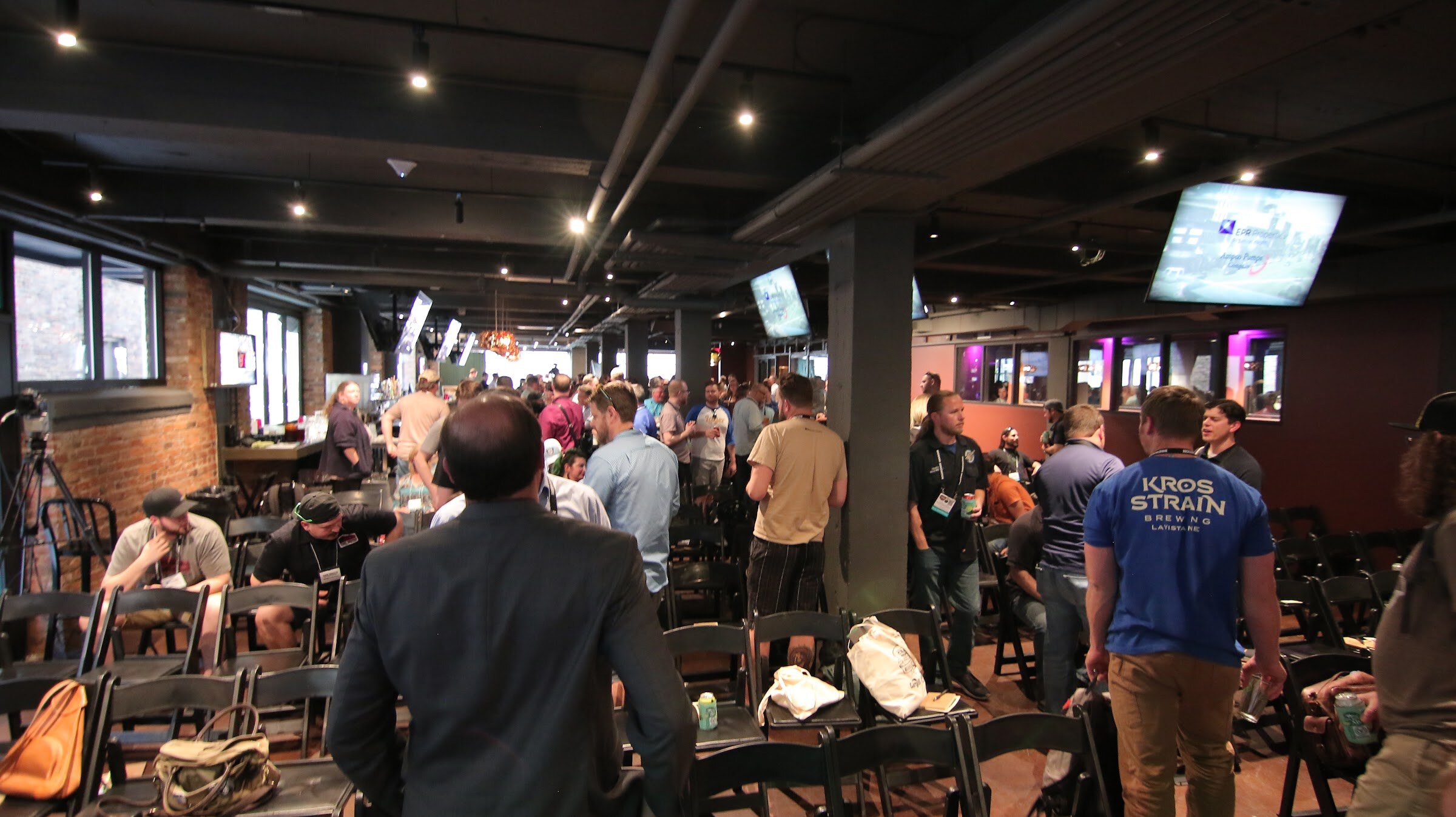 A wide view of a dark brick-walled bar where attendees stand and chat among rows of black folding chairs, with TV screens overhead and a man in a Kros Strain Brewing shirt in the foreground.
