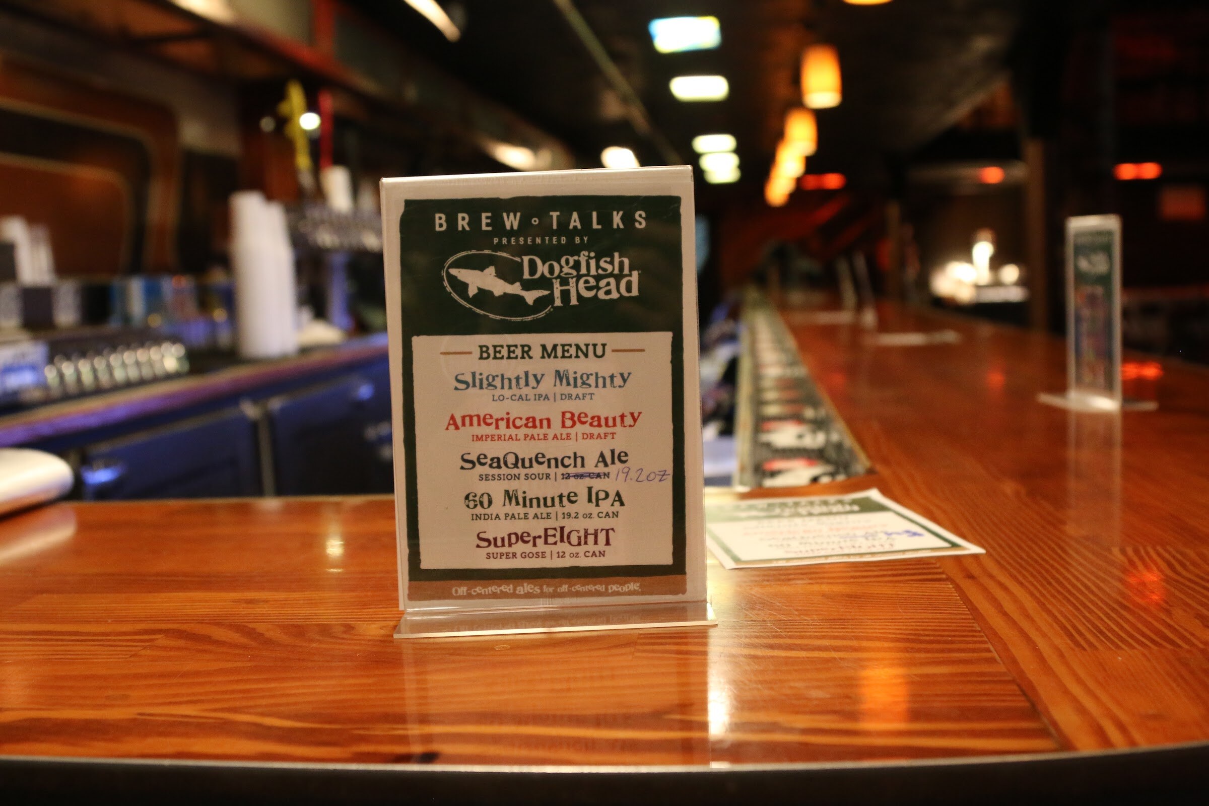 A Brew Talks beer menu card standing on a polished bar, listing Slightly Mighty, American Beauty, SeaQuench Ale, 60 Minute IPA and SuperEIGHT.