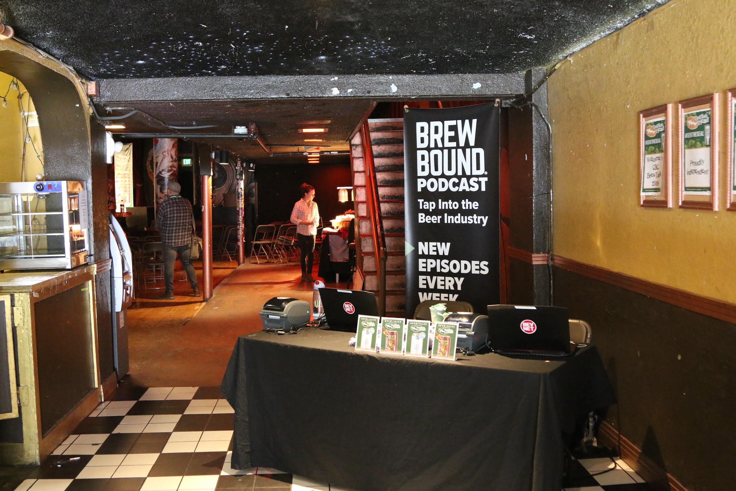 A black table with laptops and printed cards in front of a Brewbound Podcast banner in a dim hallway with a checkered floor.