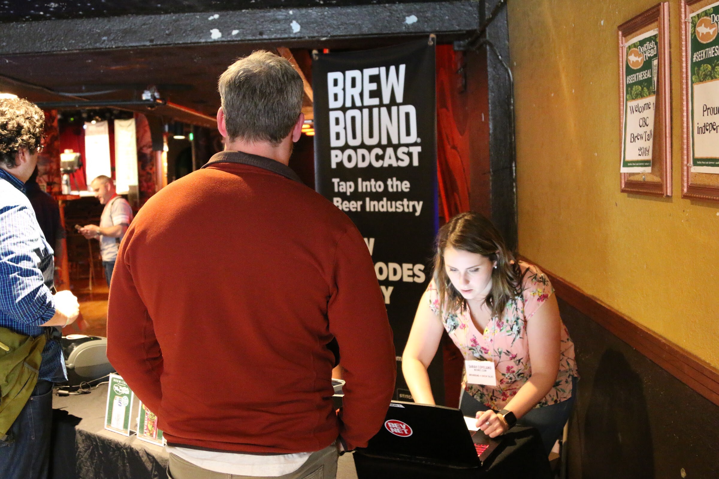A woman at a laptop checking in a man in a rust-colored sweater beside a Brewbound Podcast banner.