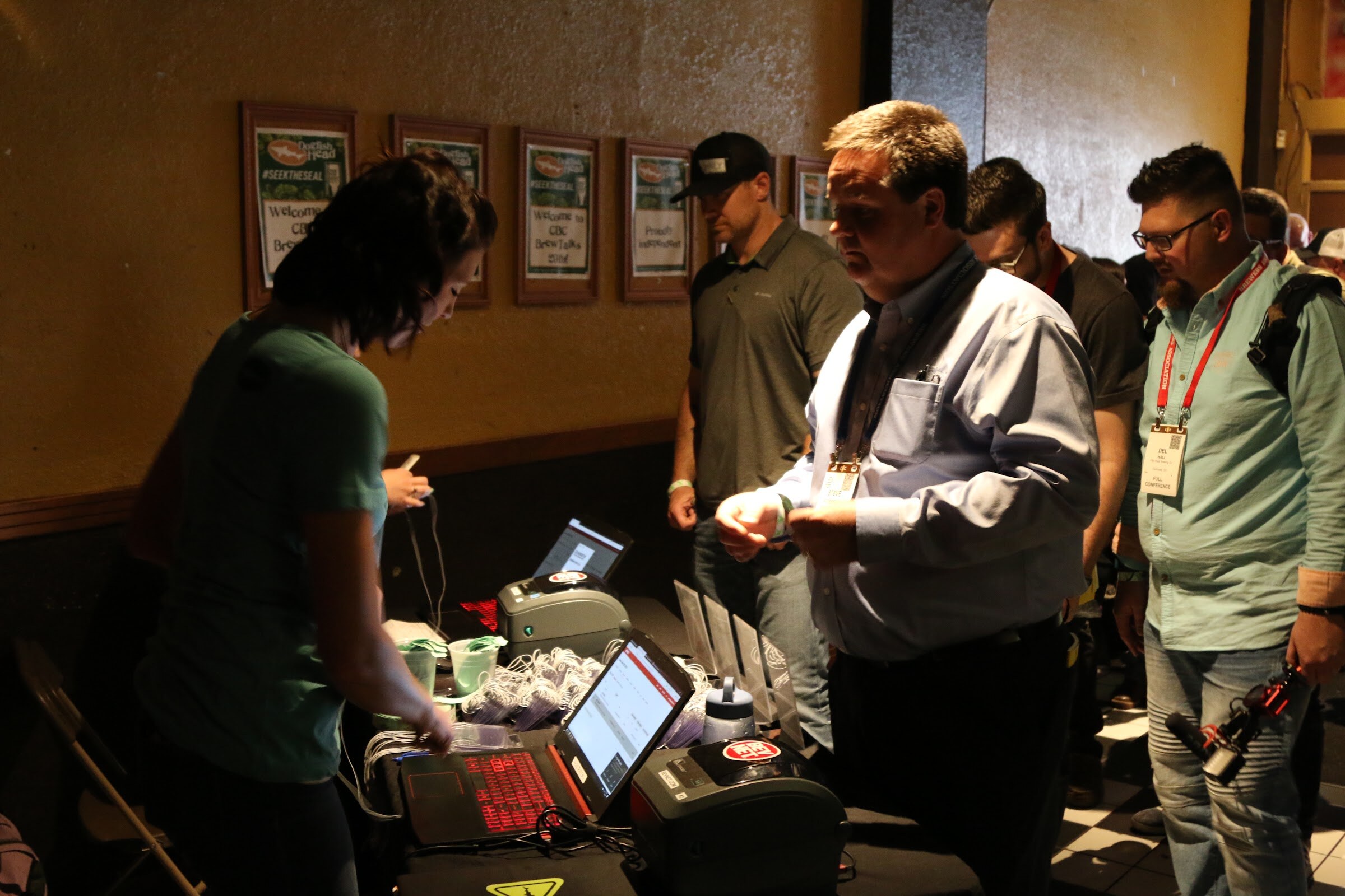 Several attendees in lanyards waiting at a table with laptops and badge printers, framed photos on the wall behind.
