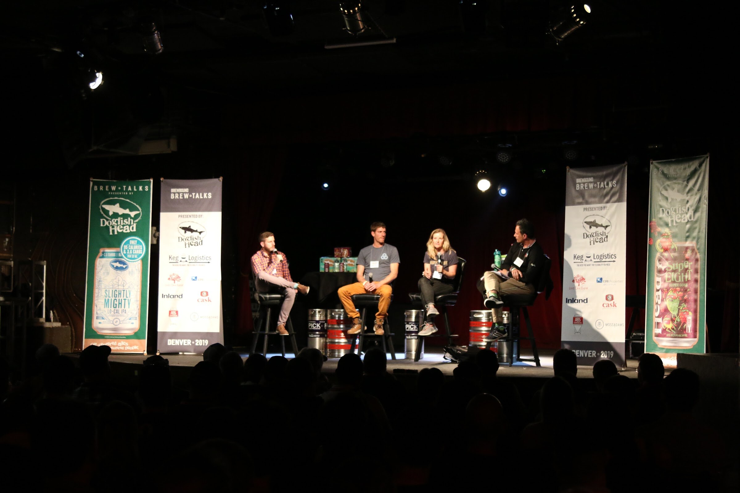 Wide shot of four people on stools on a dark stage between Dogfish Head and Brewbound Brew Talks banners, with kegs at their feet.