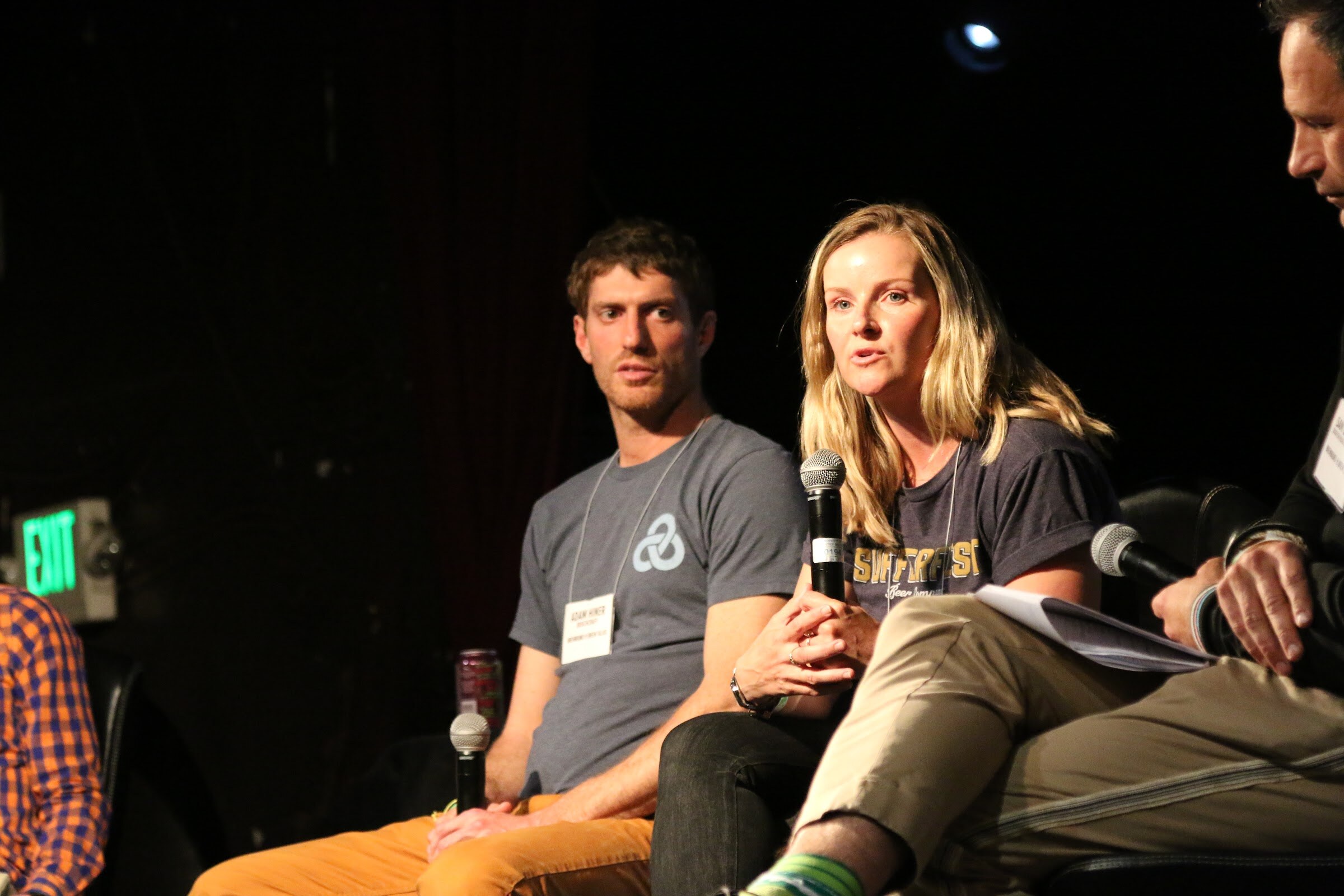 A man and a woman in gray t-shirts on stage, the woman speaking into a microphone, with an exit sign glowing behind them.