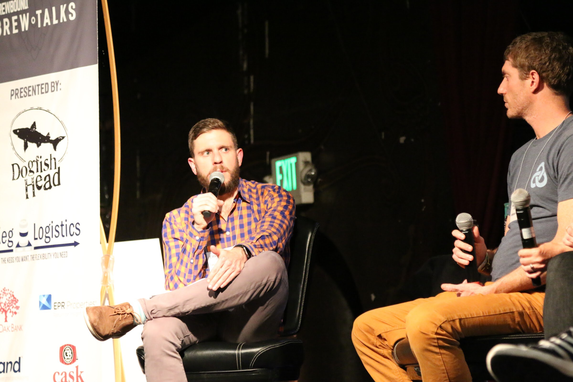 A man in an orange-and-purple checked shirt speaking into a microphone in a leather chair, a second panelist at the right, and a Presented by Dogfish Head banner at the left.