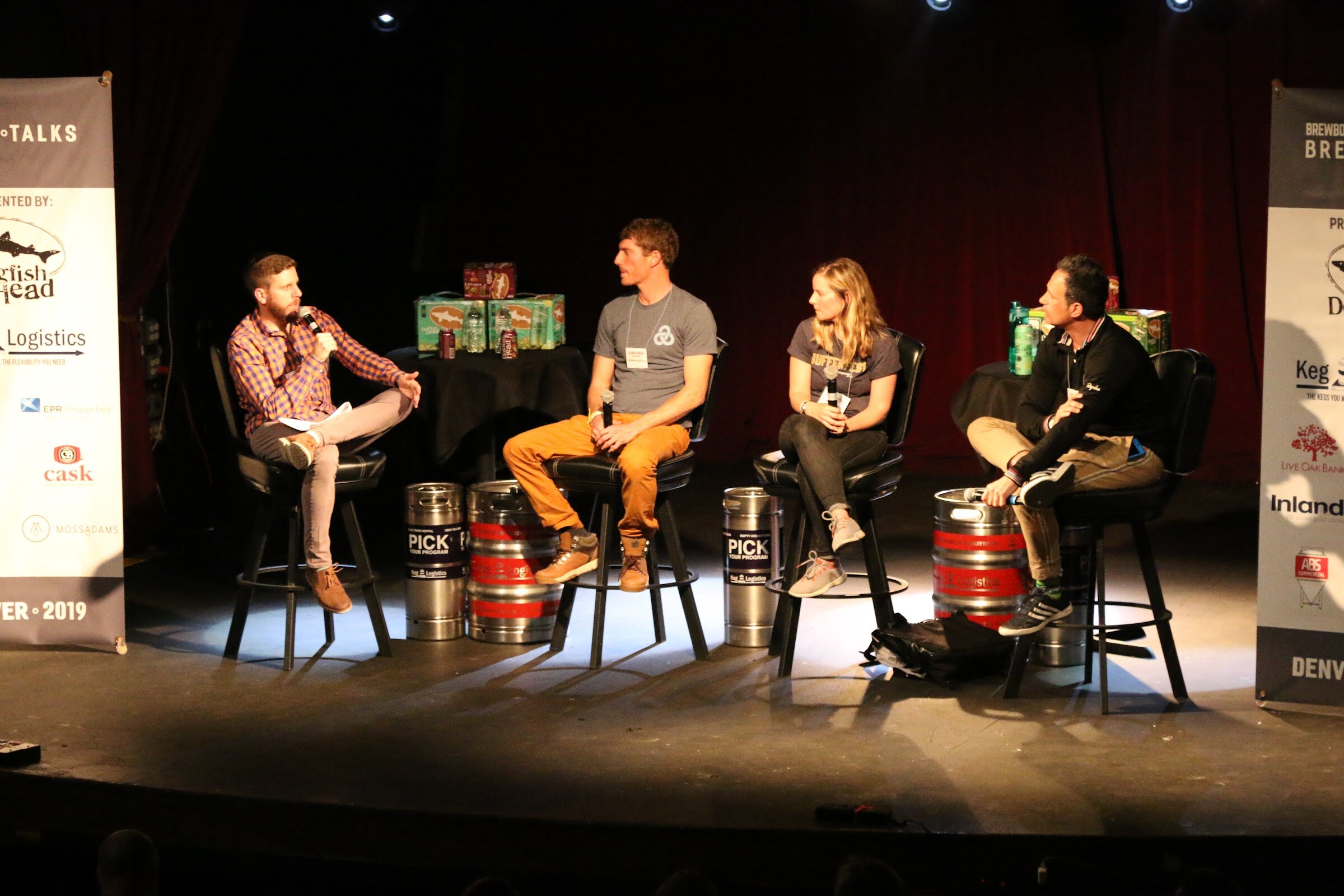 Four people on tall stools on a spotlit stage with kegs and cases of beer on tables, Brew Talks banners at the sides.