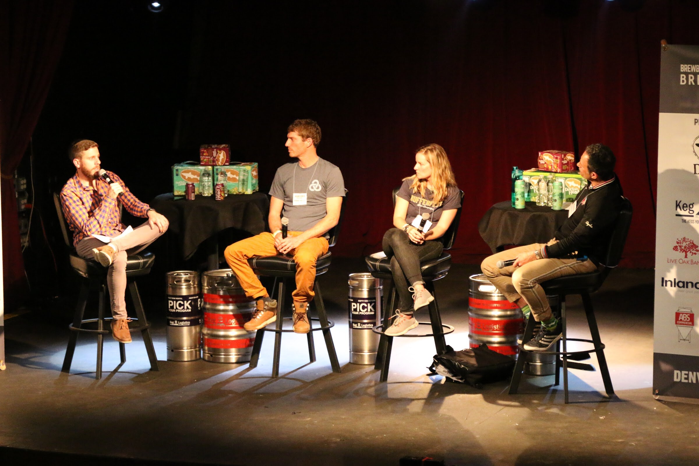 Four people seated in a row on a lit stage, the first holding a microphone, with kegs on the floor beneath them.