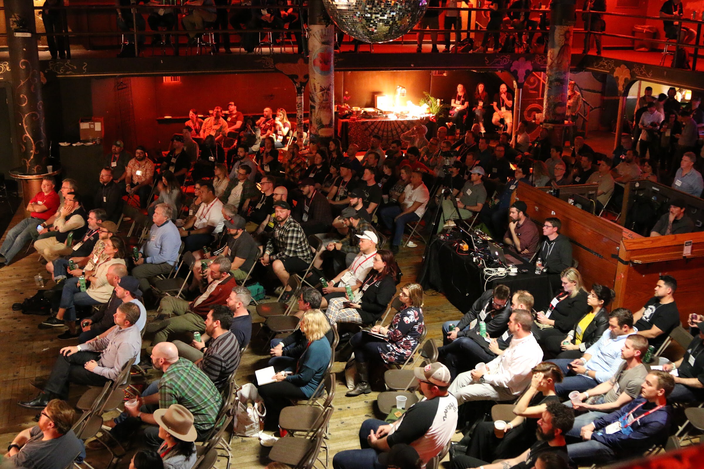 Overhead view of a packed audience seated on folding chairs in a red-lit music hall with a disco ball above.