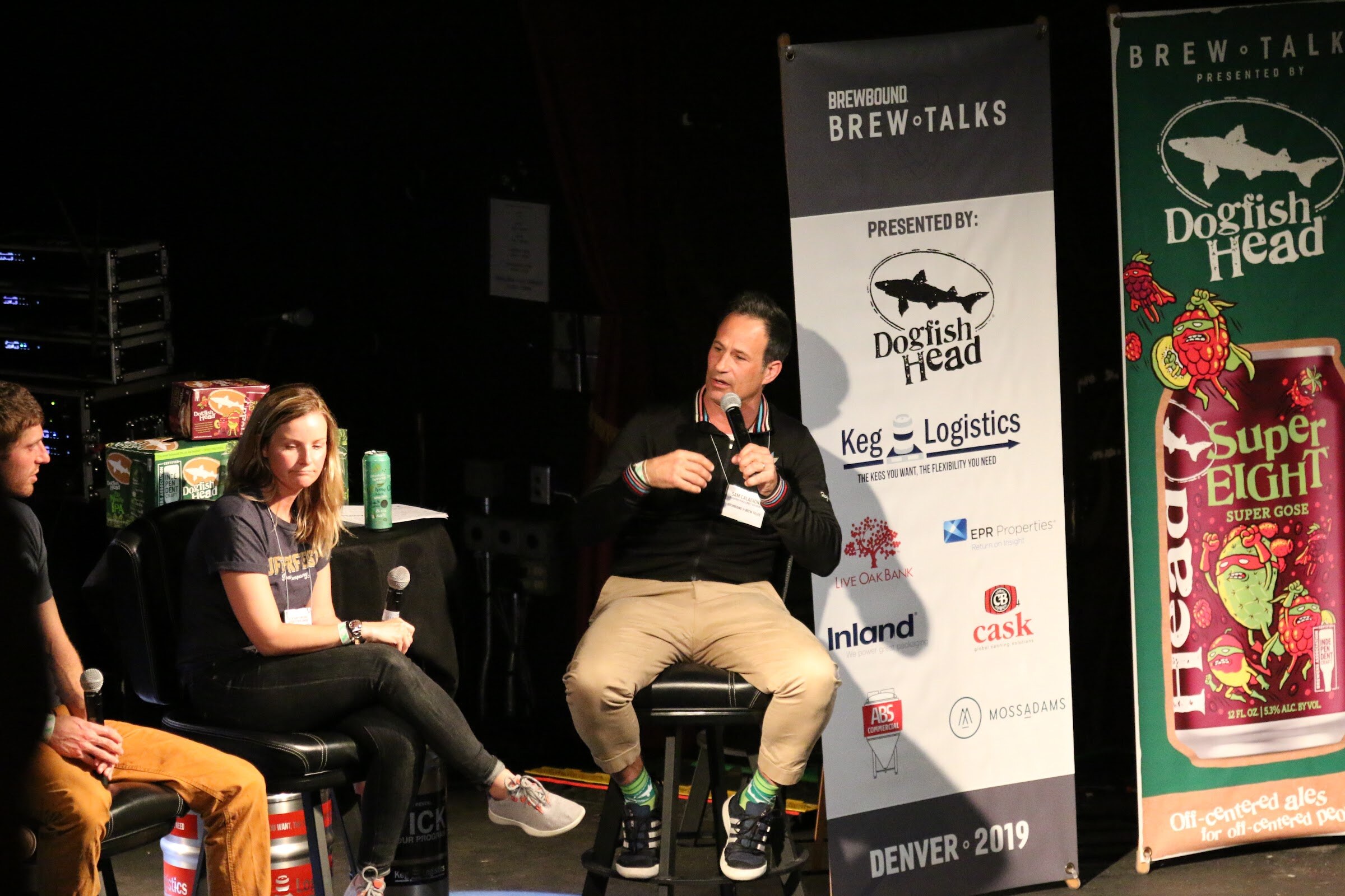 A man on a stool speaking into a microphone and a woman seated beside him, next to banners for Brewbound Brew Talks and Dogfish Head SuperEIGHT.