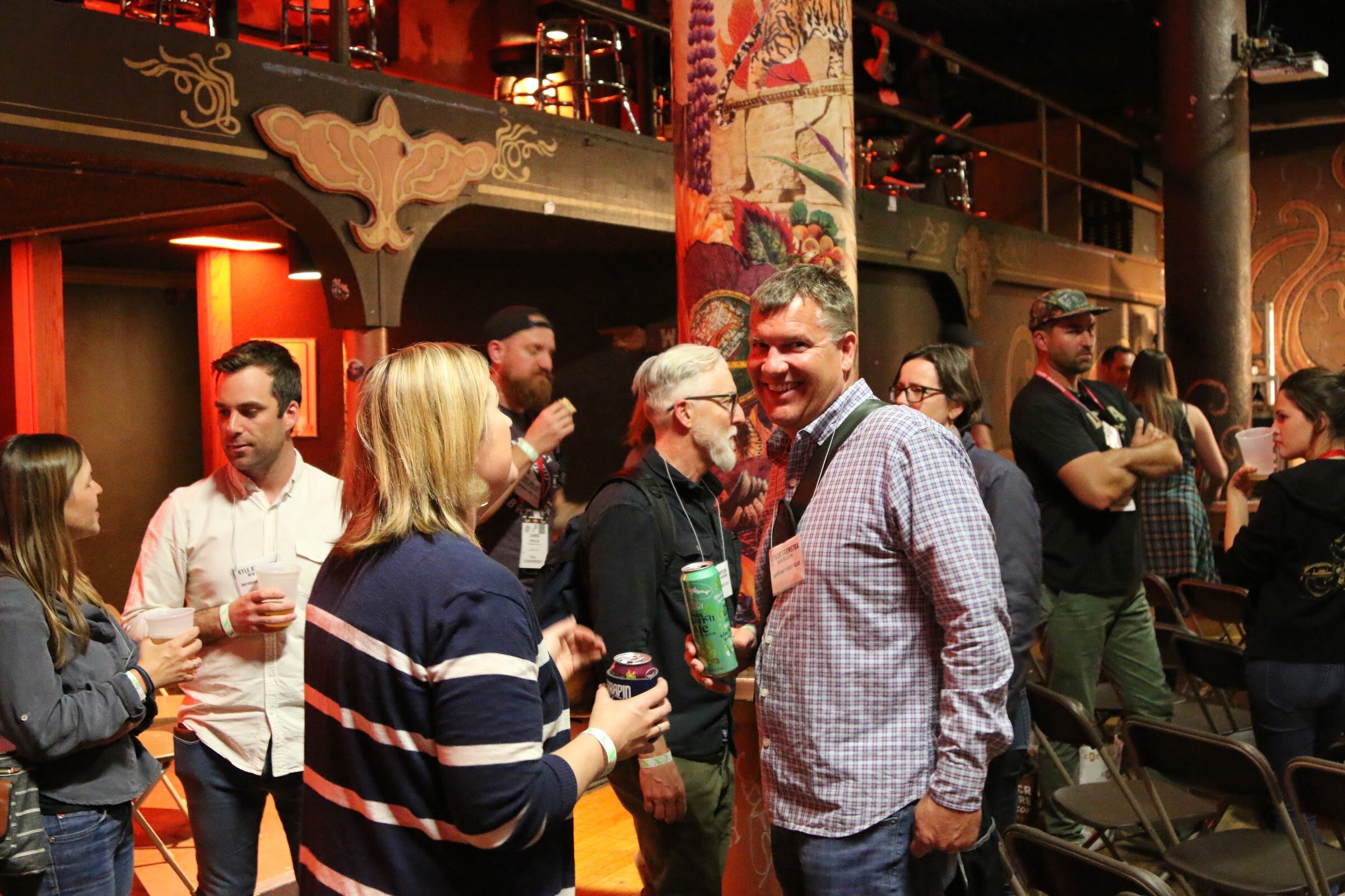 A group of attendees with lanyards and drinks talking in a venue with ornate painted columns.