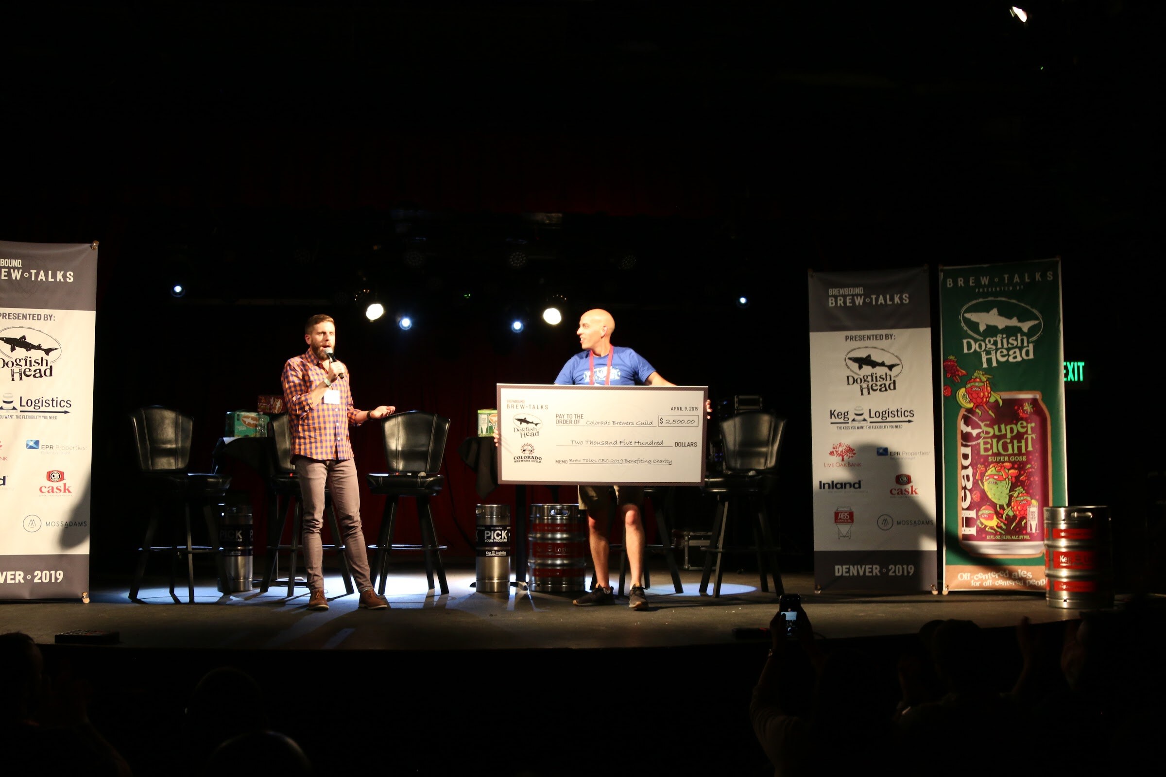 A spotlit stage with two people holding an oversized check between Brew Talks banners.