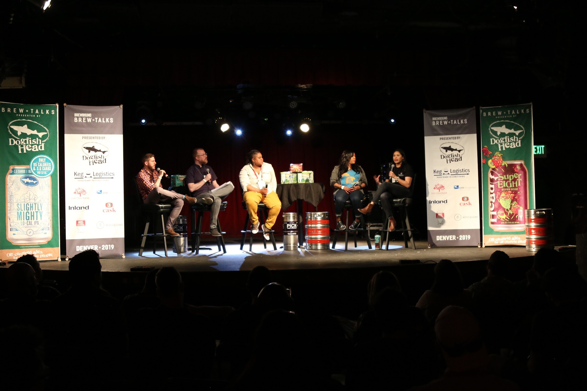 Wide shot of five people seated on stools on a stage with Brew Talks banners and kegs at their feet.