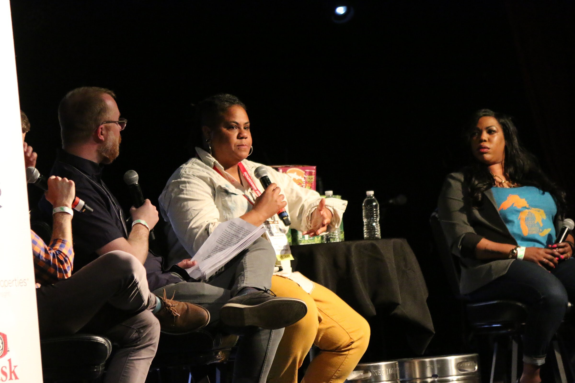 Several seated panelists on stage, one holding papers and a microphone, another in a blue shirt with a graphic.
