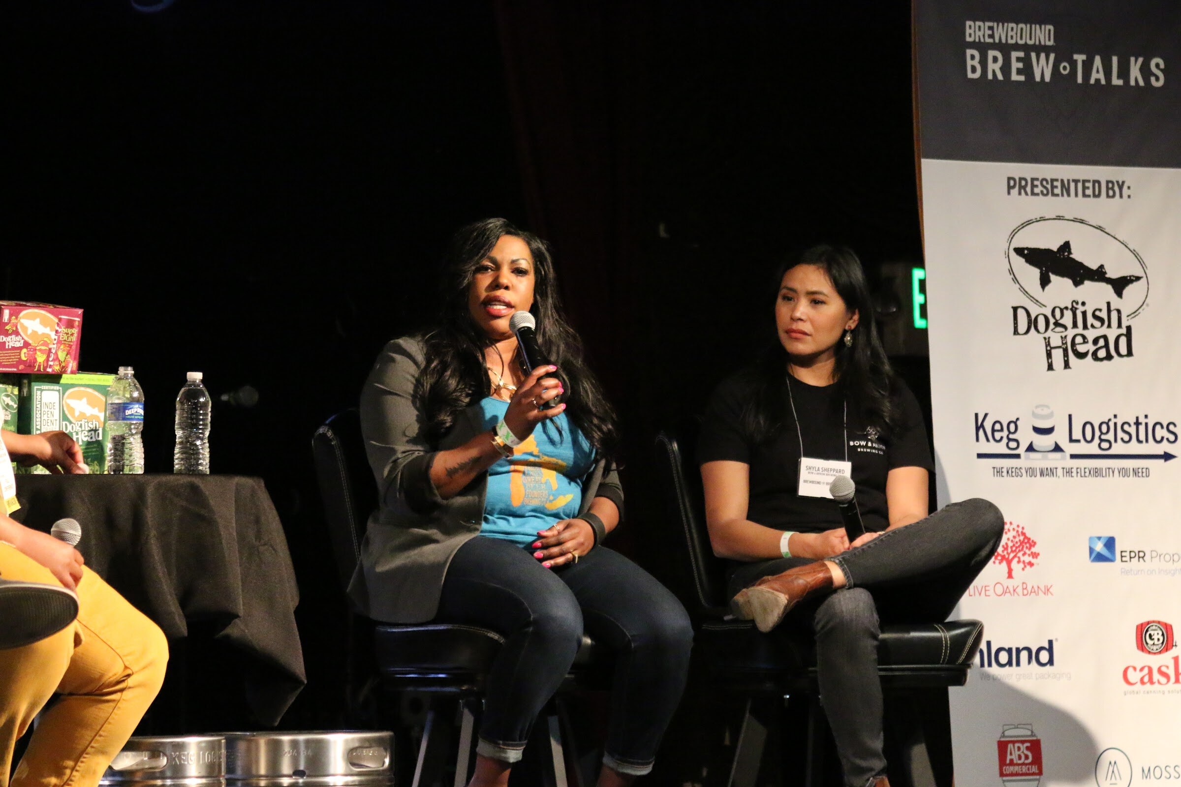 Two women seated on stage, one speaking into a microphone, next to a Brew Talks banner listing sponsors.