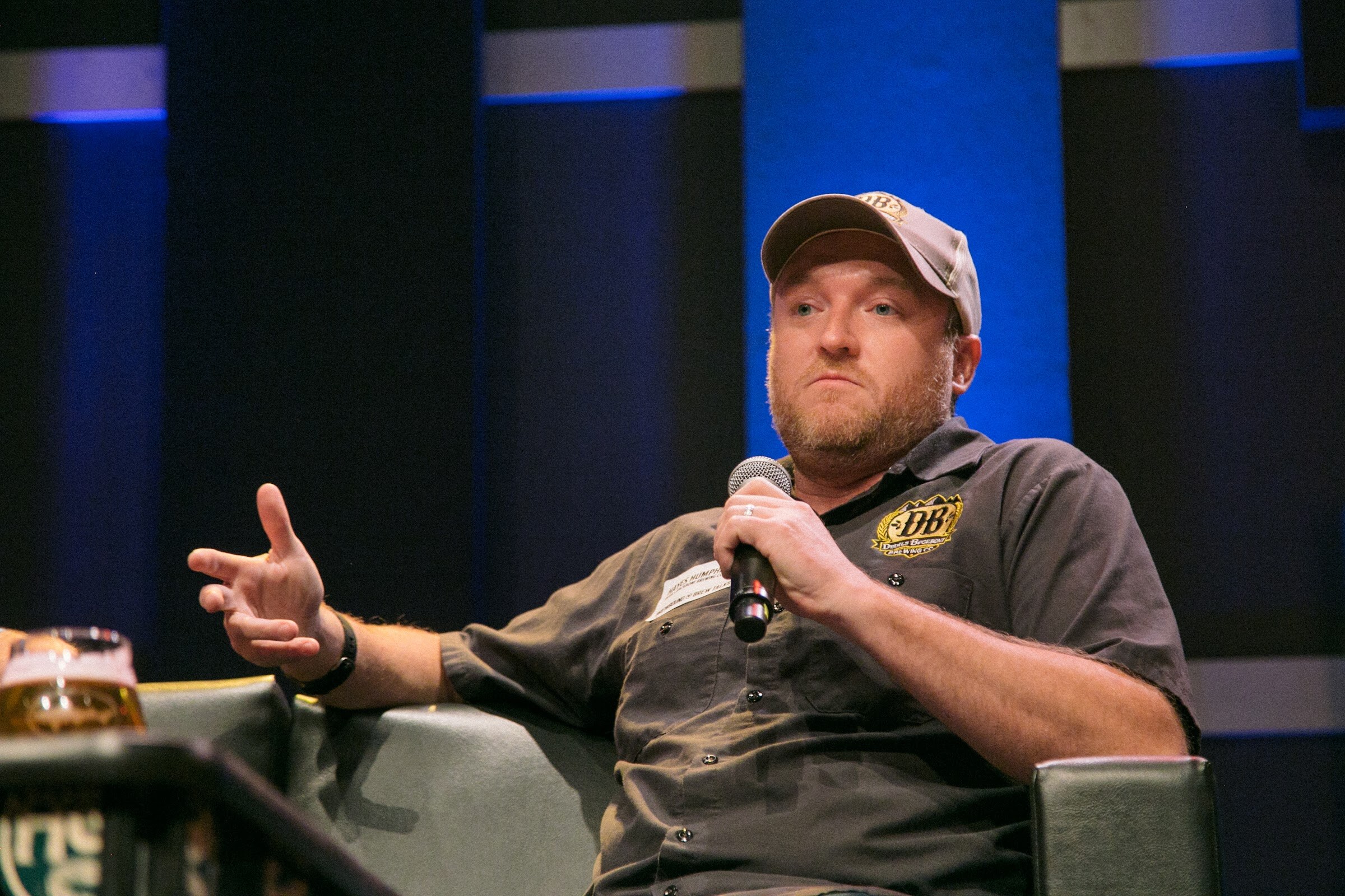 A man in a cap and dark gray shirt holds a microphone and extends his free hand from an armchair.