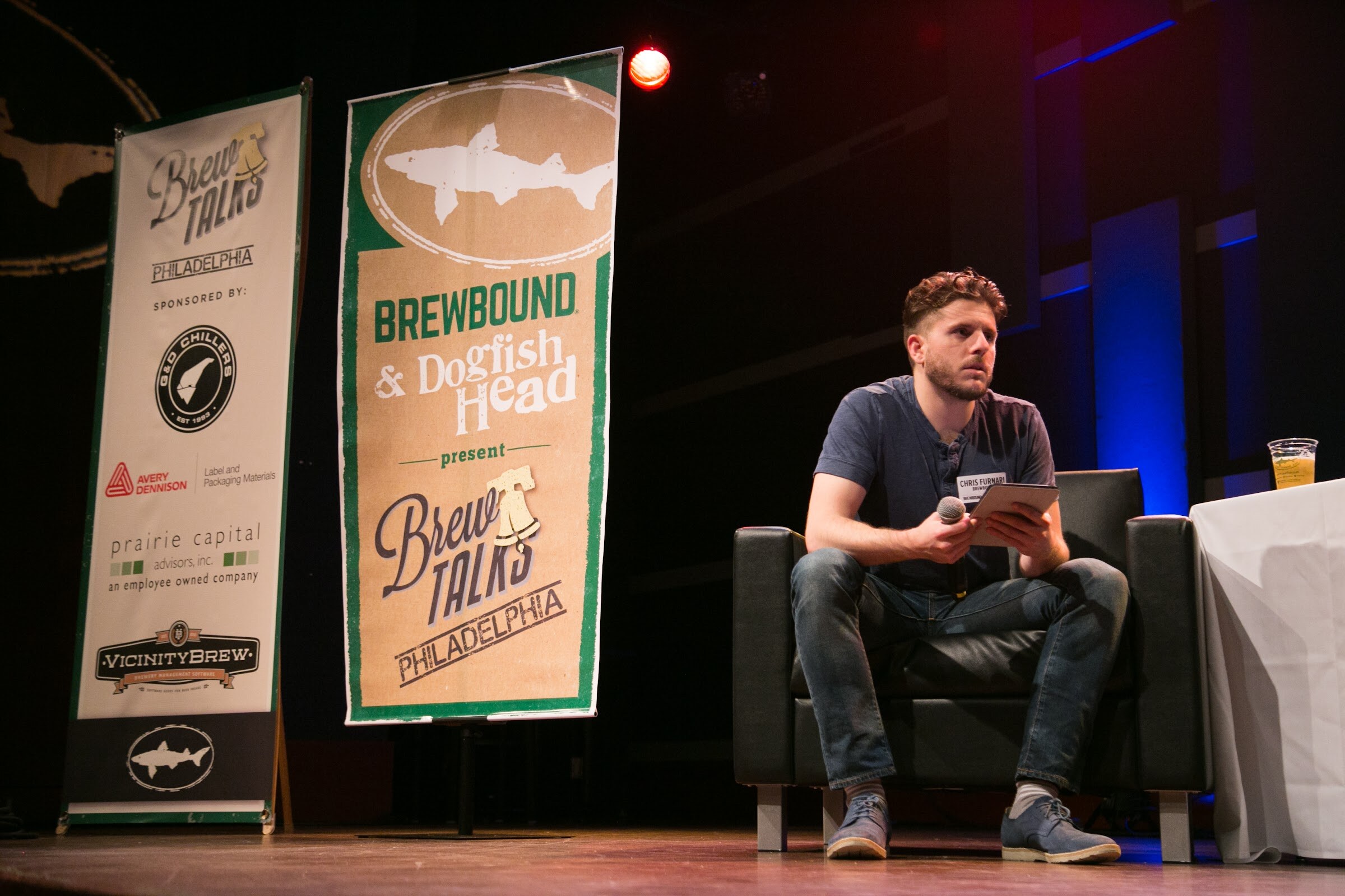 A man in a navy shirt sits with a tablet and microphone between a table and tall Brewbound and Dogfish Head banners.