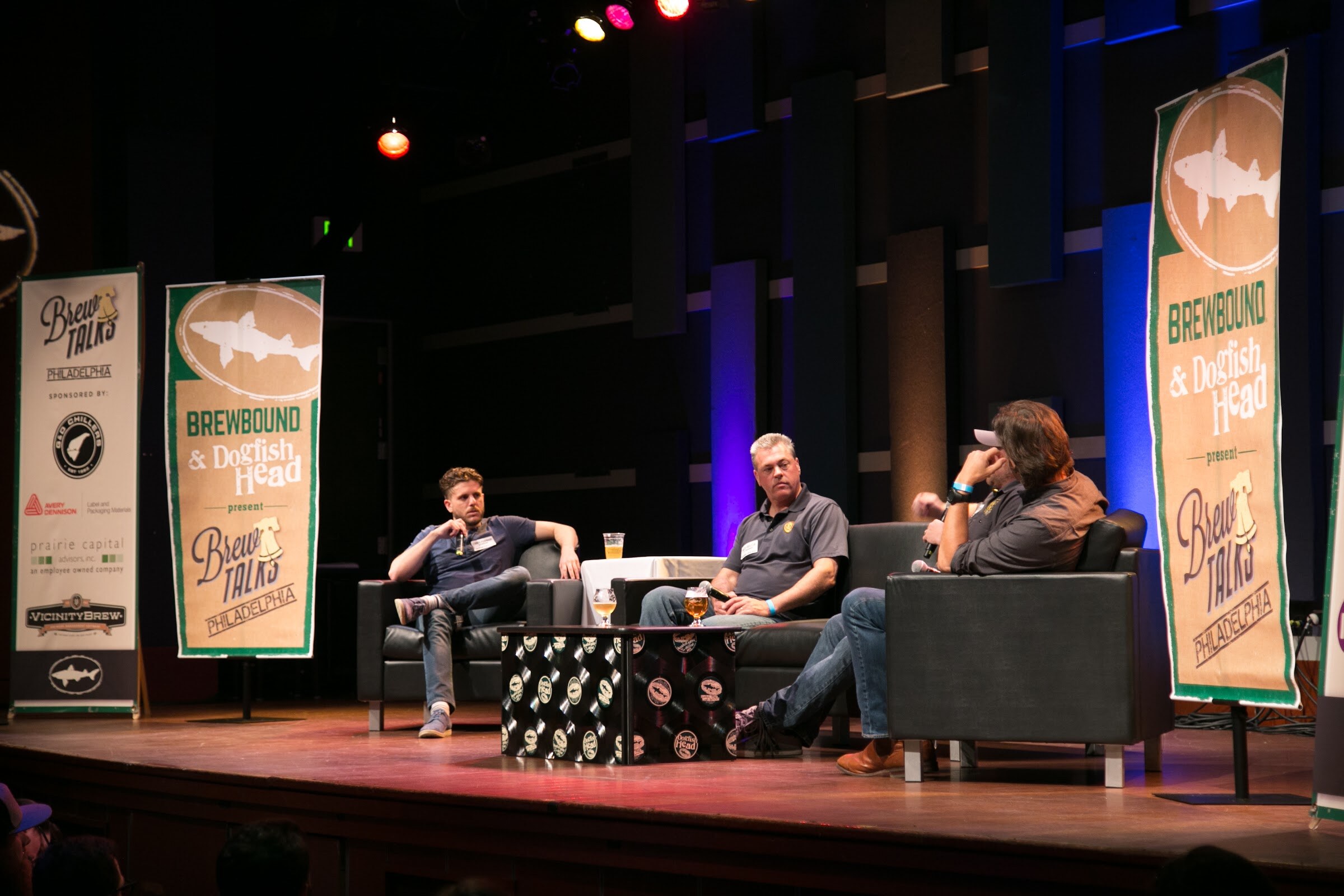 Seated men look across a stage furnished with a sofa, armchairs and a record-decorated table between event banners.