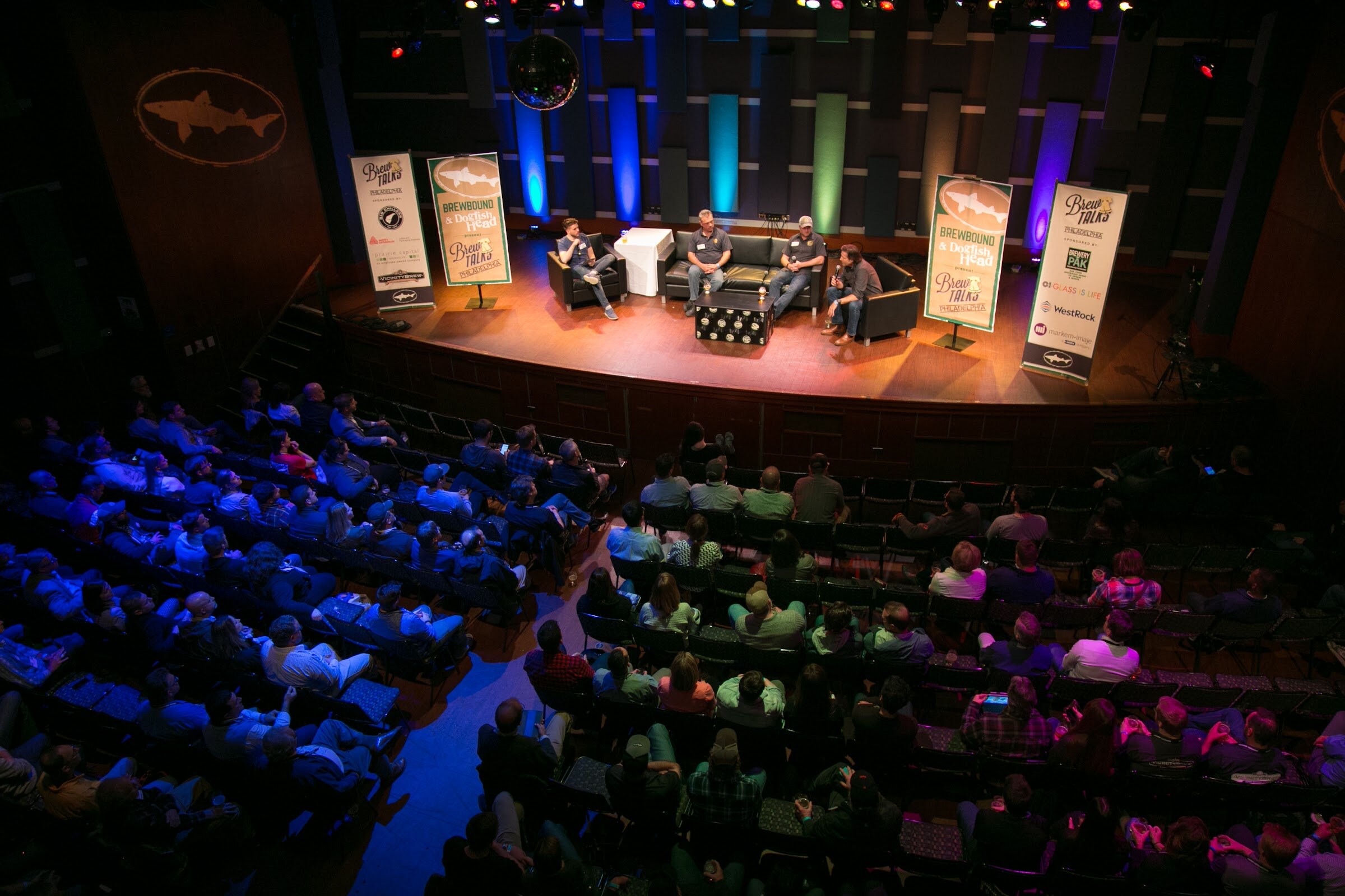 An elevated view shows rows of seated attendees facing four men on a warmly lit stage flanked by event banners.