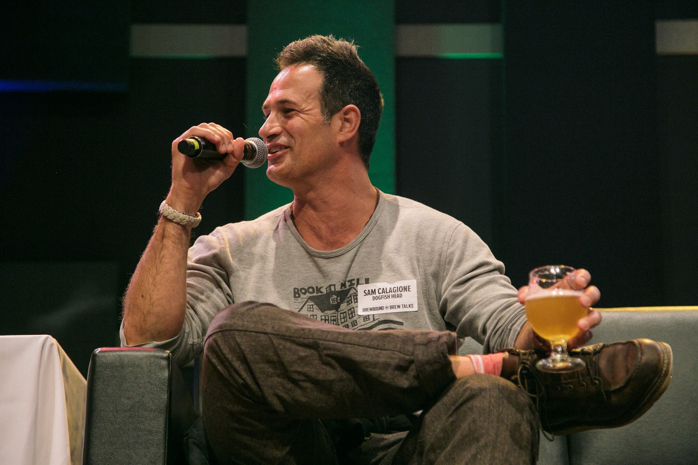 A smiling man in a gray long-sleeve shirt sits cross-legged with a microphone in one hand and a beer glass in the other.