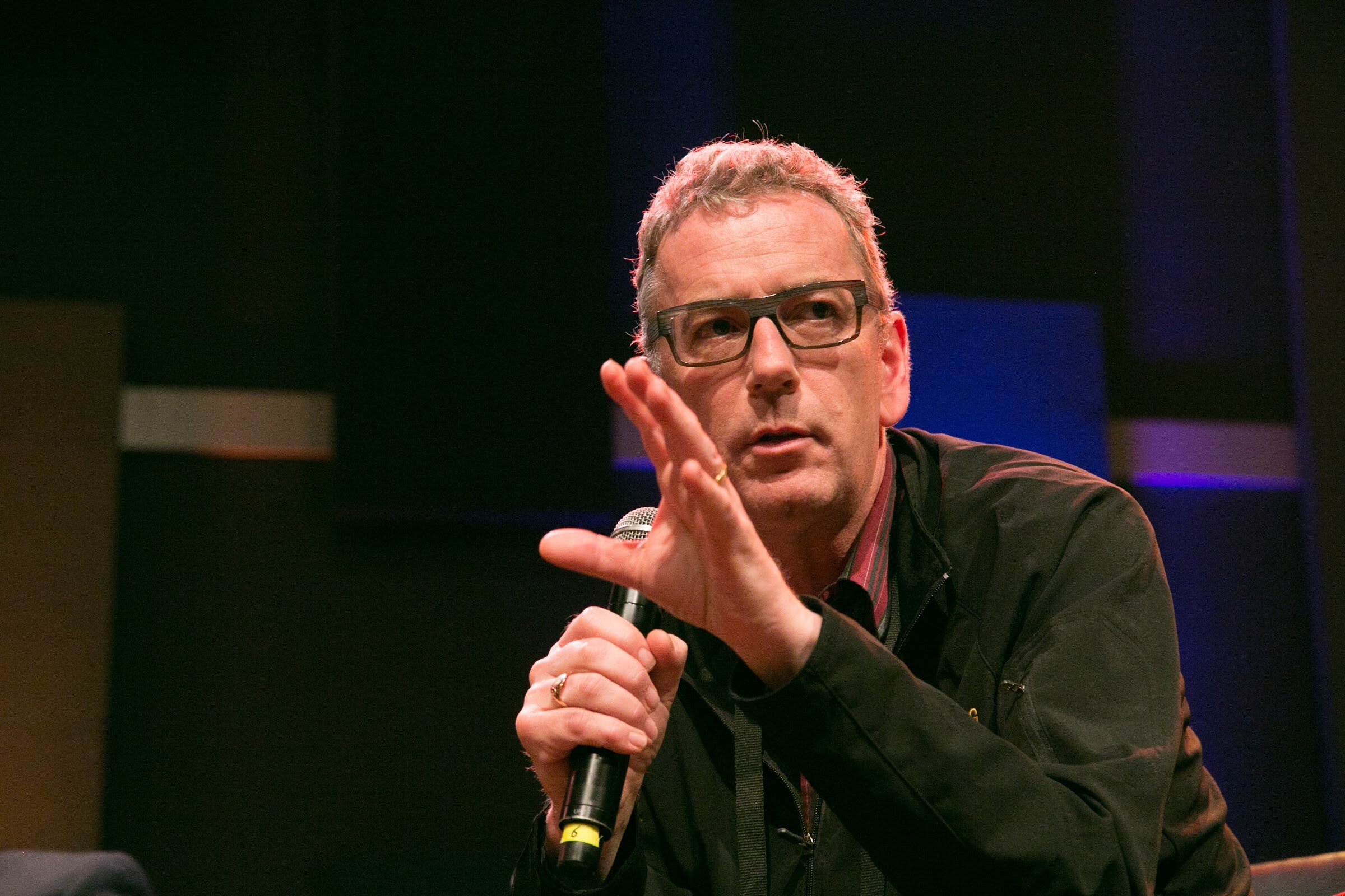 A man wearing glasses and a black shirt holds a microphone and raises his other hand against a dark blue-lit background.